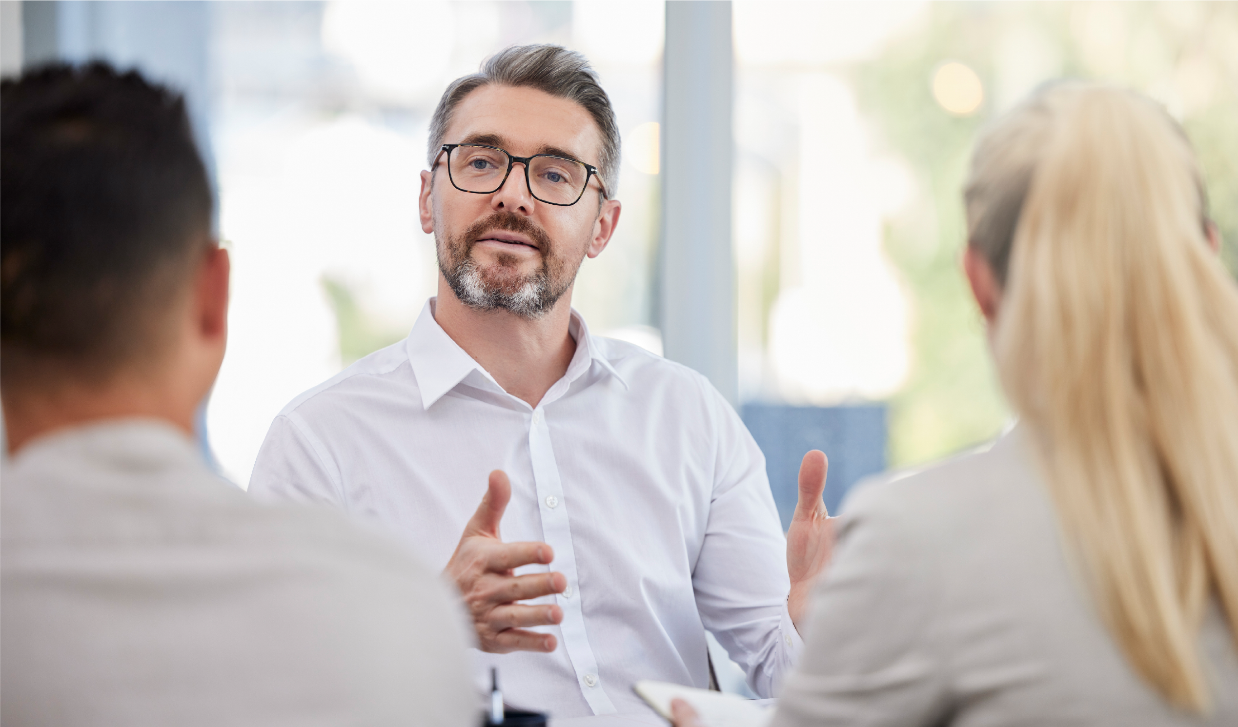 Man in white shirt and glasses speaking to two people in a bright office setting.