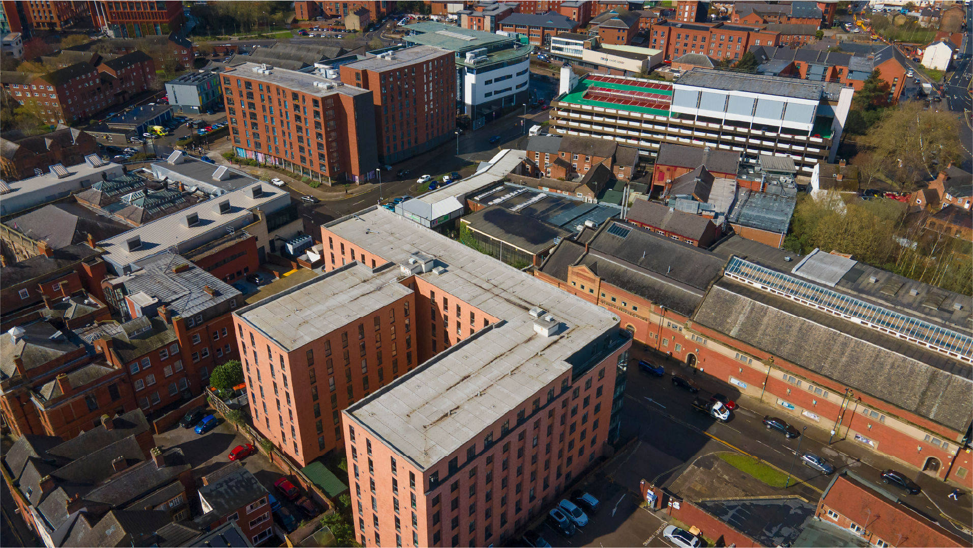 Aerial view of a city block with red-brick buildings, streets, and parked cars on a sunny day.