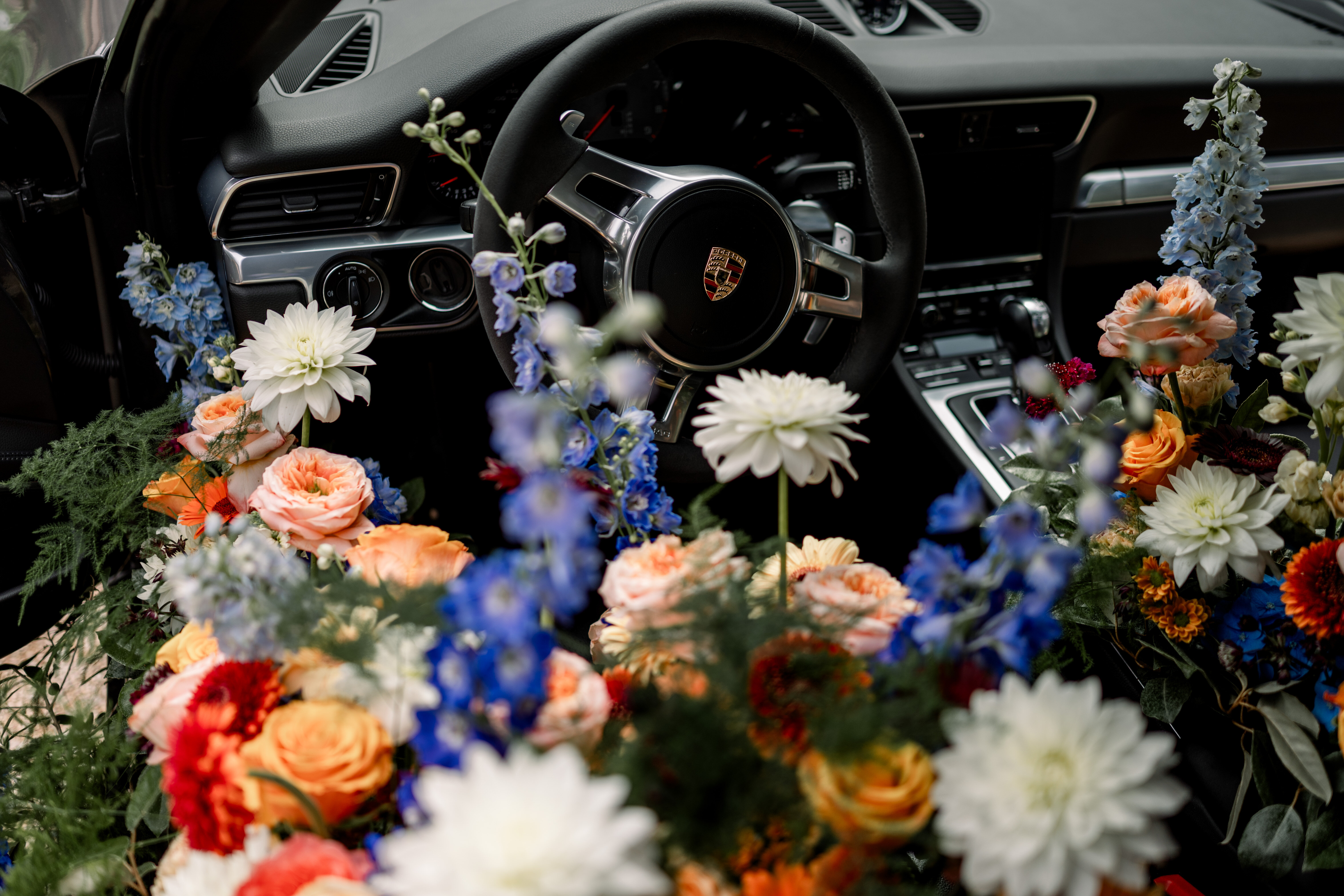 Intérieur de voiture de luxe avec volant Porsche entouré de nombreuses fleurs colorées, notamment des roses orange, des dahlias blancs et des fleurs bleues.