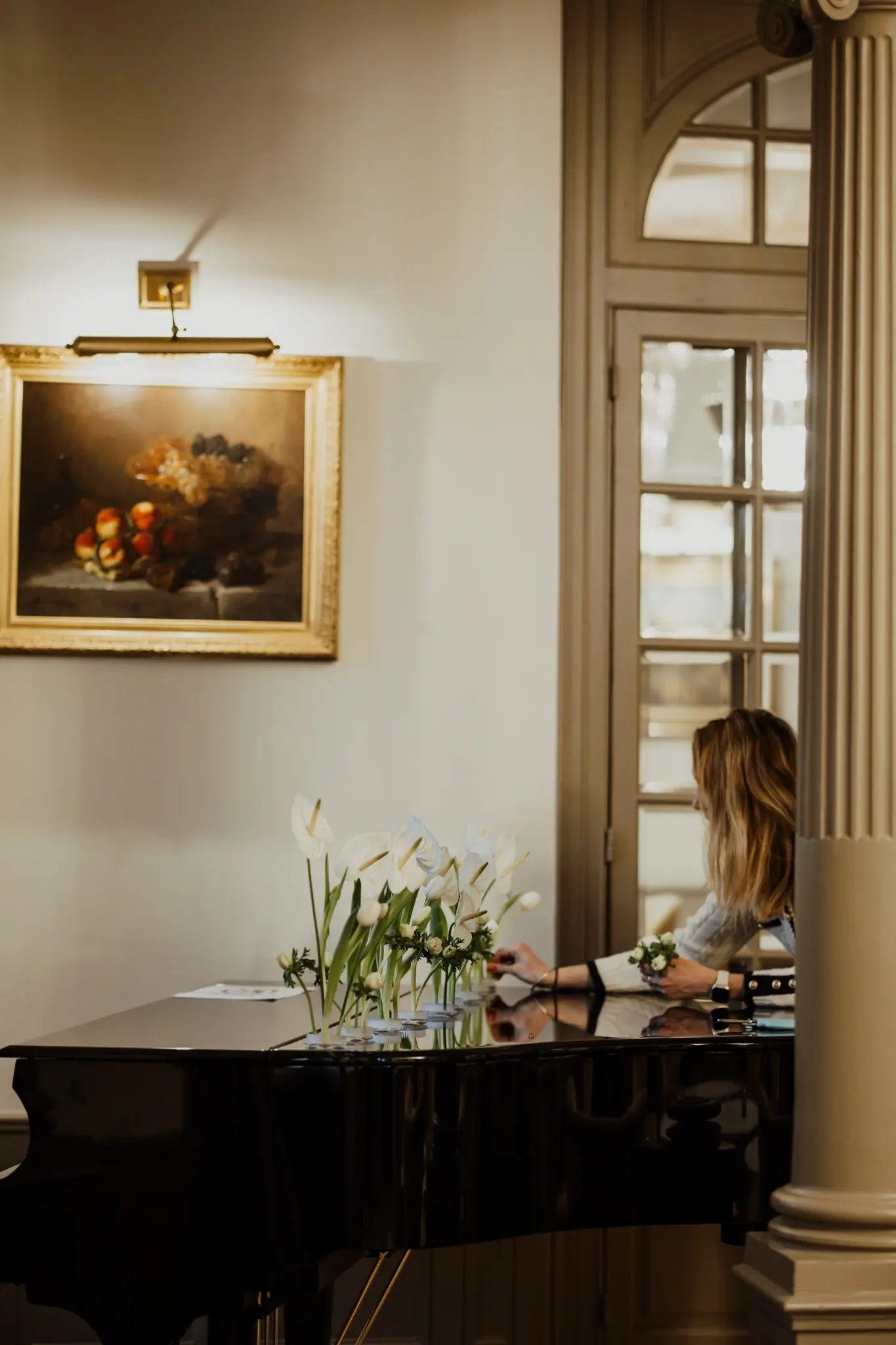 Une femme arrange des fleurs blanches sur un piano à queue noir dans une pièce élégante avec une grande porte vitrée et un tableau encadré accroché au mur.