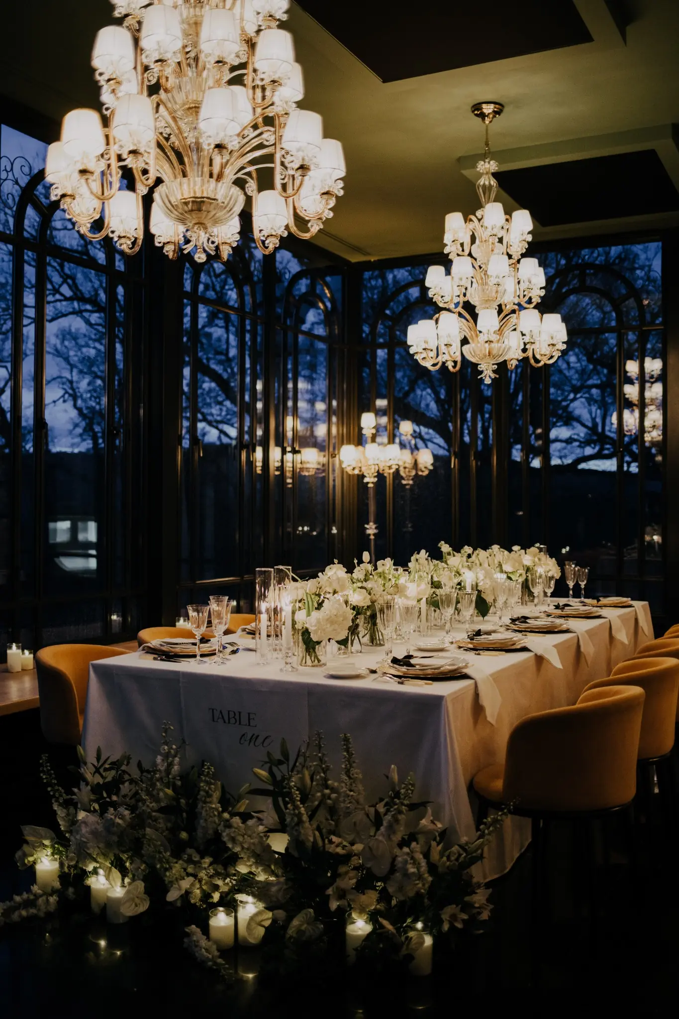 Une table élégamment dressée avec une nappe blanche portant l'inscription Table One, décorée de bouquets de fleurs blanches et de bougies allumées sur le sol à l'avant, entourée de fauteuils jaunes dans une pièce aux grandes fenêtres en verre réfléchissant des lustres lumineux.