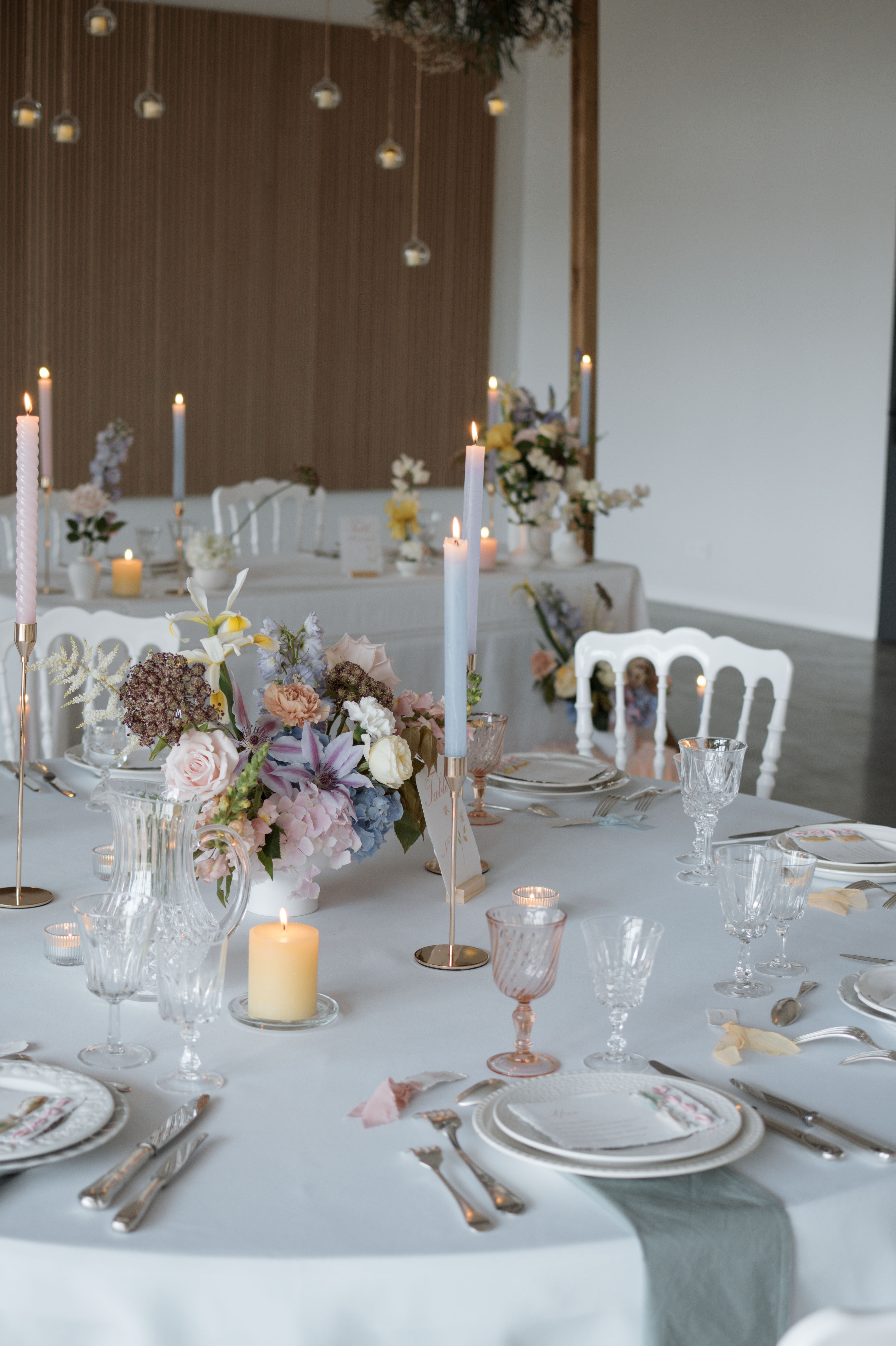 Table élégamment dressée avec une nappe blanche, des bougies allumées, un centre de table floral aux teintes pastel et des verres en cristal, prête pour un repas festif.