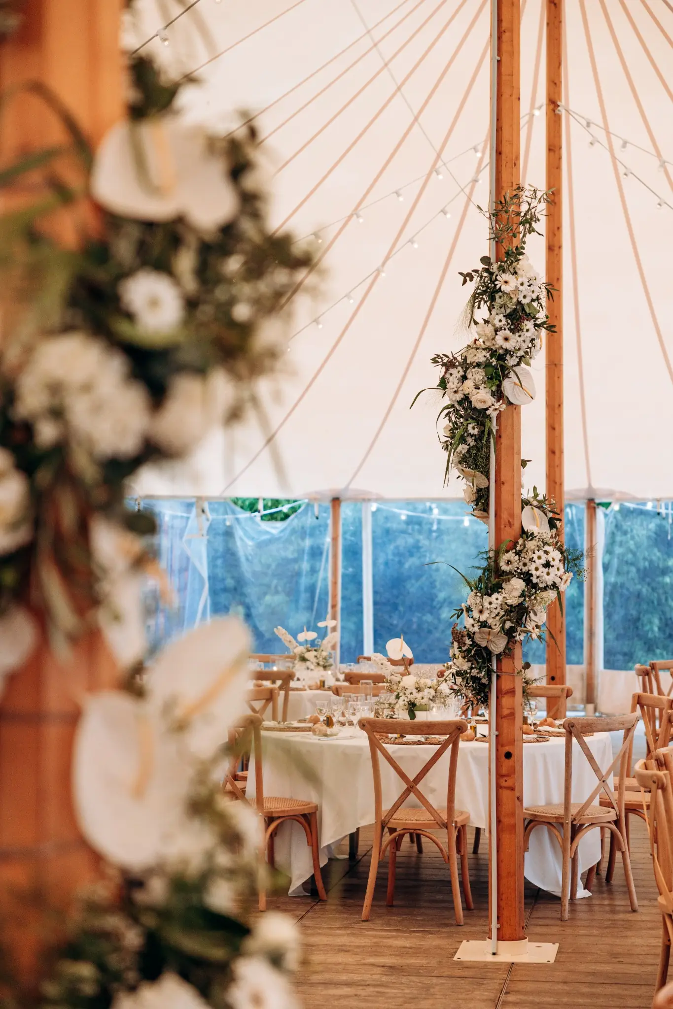 Intérieur d'une grande tente décorée pour un événement, avec des tables rondes couvertes de nappes blanches, des chaises en bois naturel, et des poteaux ornés de guirlandes florales blanches et vertes, sous un toit crème avec des guirlandes lumineuses suspendues.