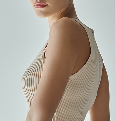 A close-up of a woman’s upper body, featuring her shoulder and part of her arm, wearing a fitted beige ribbed tank top.