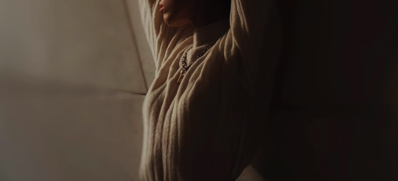 A person wearing a light-colored sweater, with their arms raised and leaning against a wall, partially side-lit to create a dramatic effect.