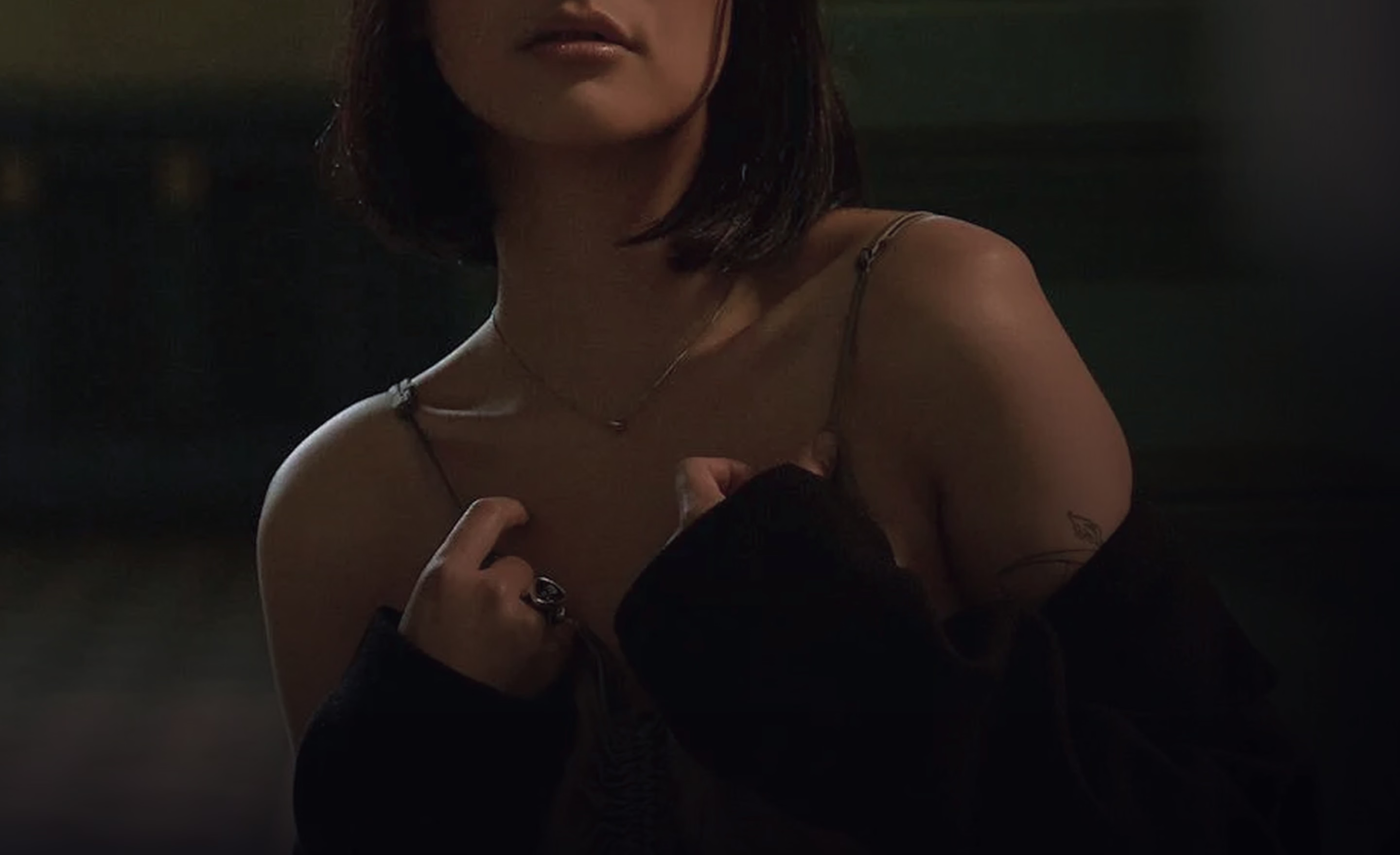 A person with short dark hair wearing a black oversized sweater, revealing a delicate chain necklace and showcasing skin on their shoulder. They are partially turned with a focused expression.