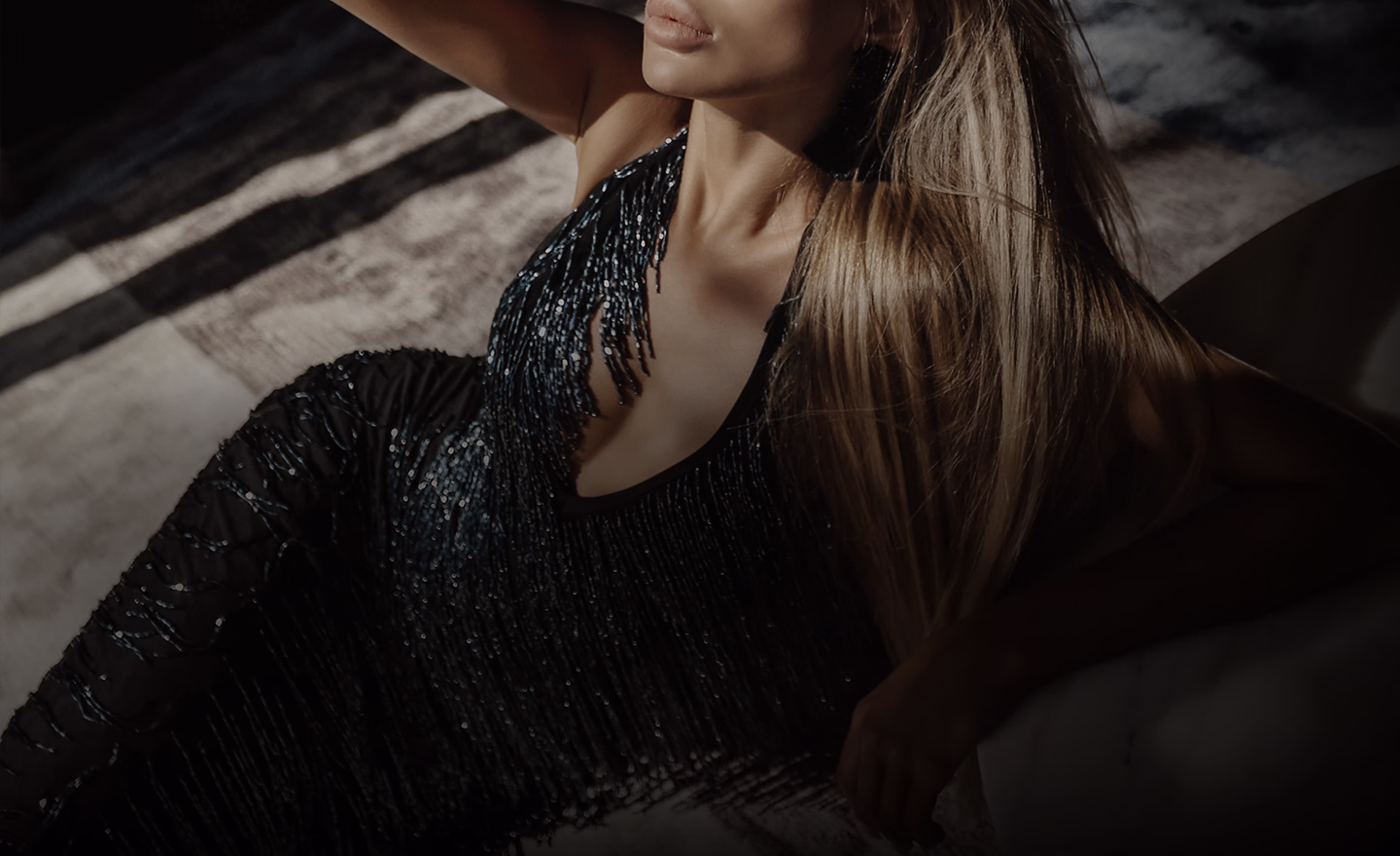 A woman in a sparkling black dress with fringe detailing, sitting and posing with one hand raised, showing long hair and a subtle smile.