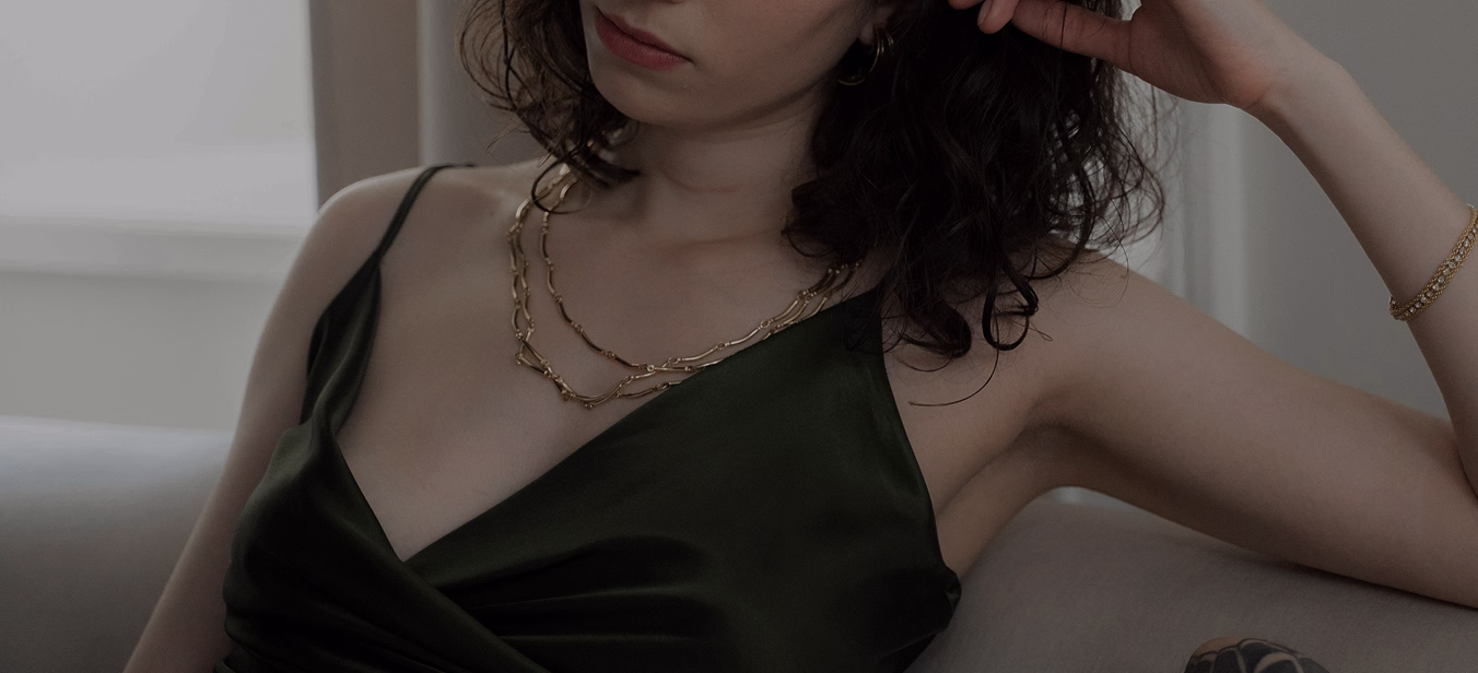 A woman wearing a green satin dress with a deep neckline, adorned with two gold chain necklaces, sitting on a couch.