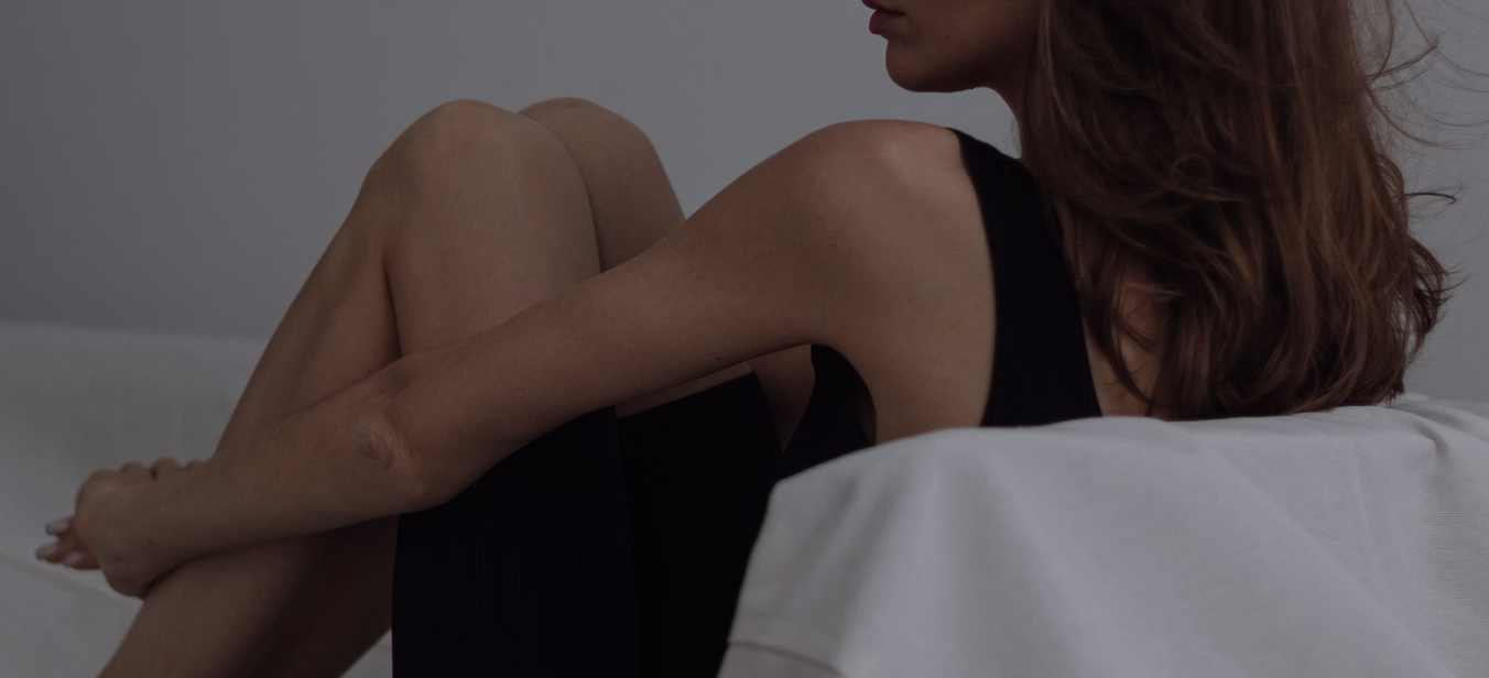 A side profile of a woman seated with her legs pulled close to her body, wearing a black sleeveless top, with soft lighting accentuating her features.