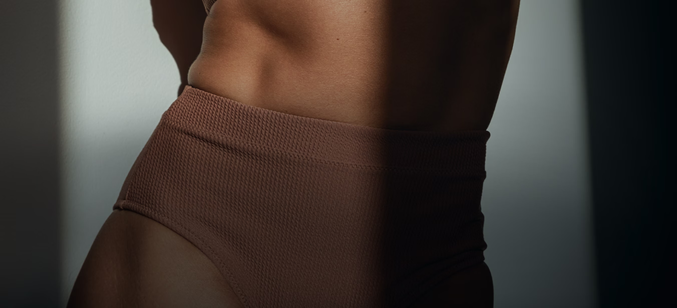 A close-up of a partially illuminated torso wearing high-waisted brown swimwear, highlighting contours and textures.