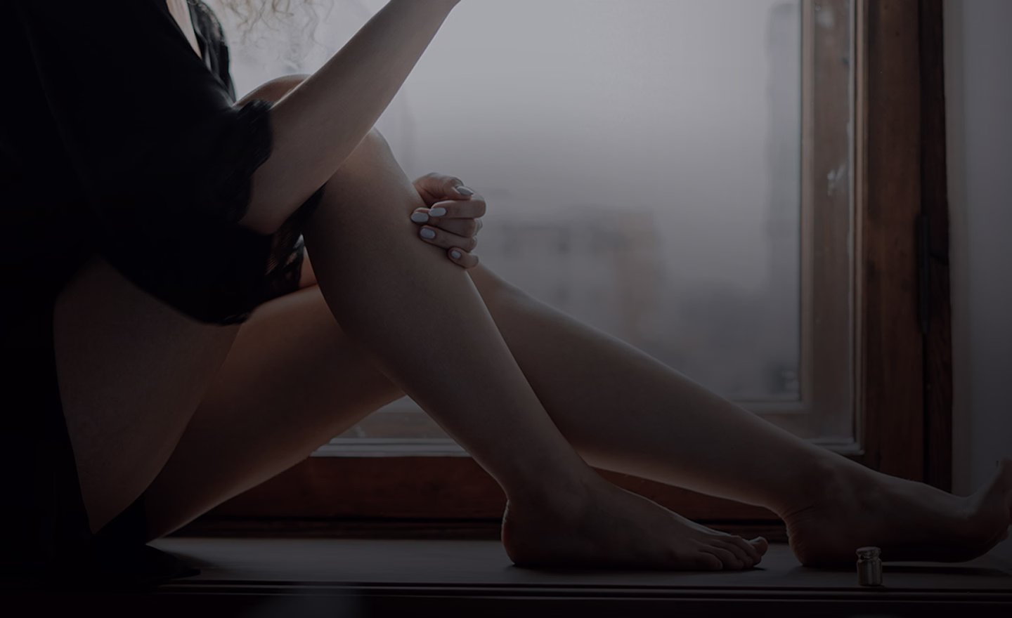 A close-up view of a woman's bare legs resting on a windowsill, with one hand gently holding the knee and another arm partially visible. The woman has manicured nails.