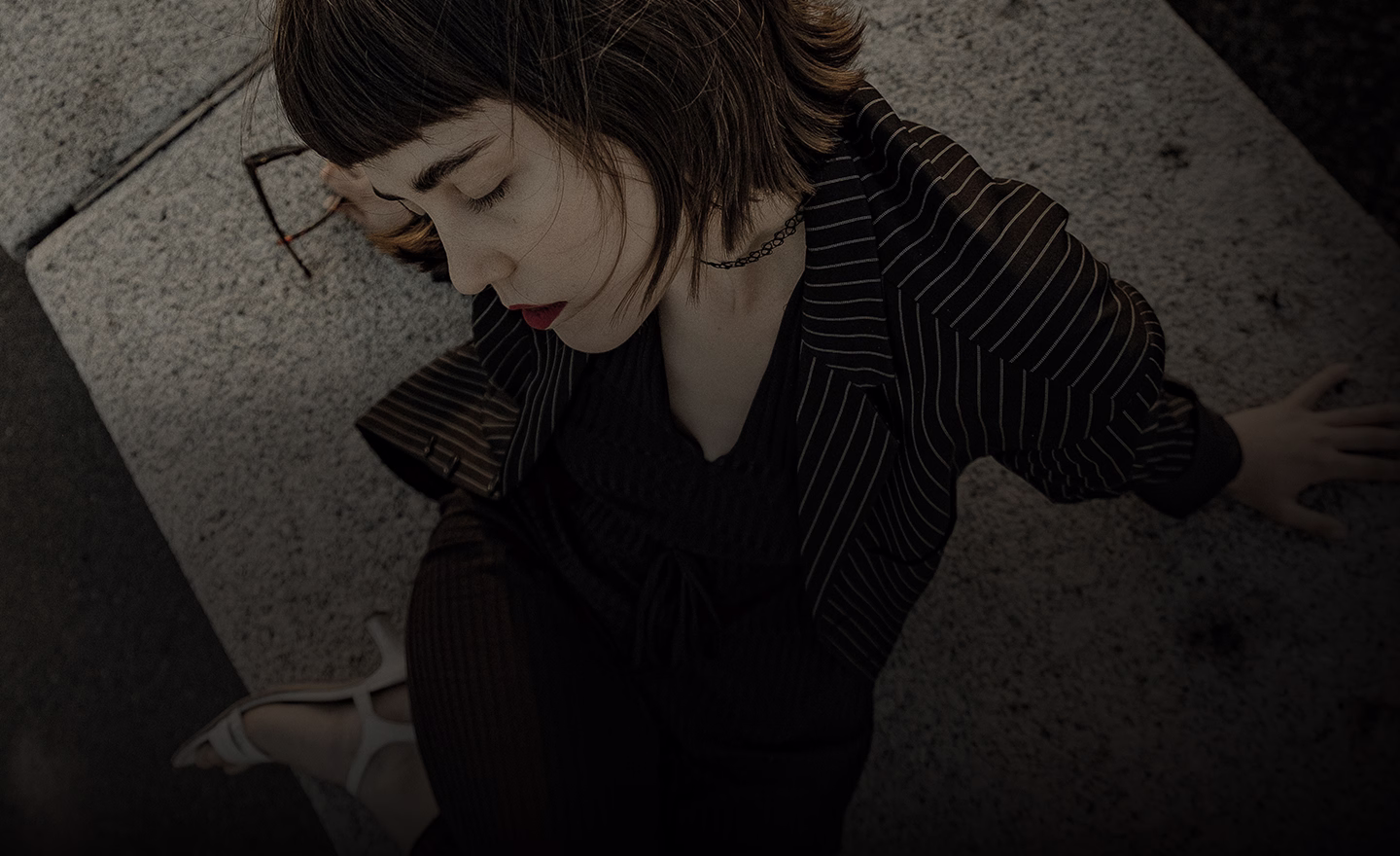 A woman in a striped suit, sitting with her eyes closed, wearing a choker necklace and posing in a thoughtful manner.