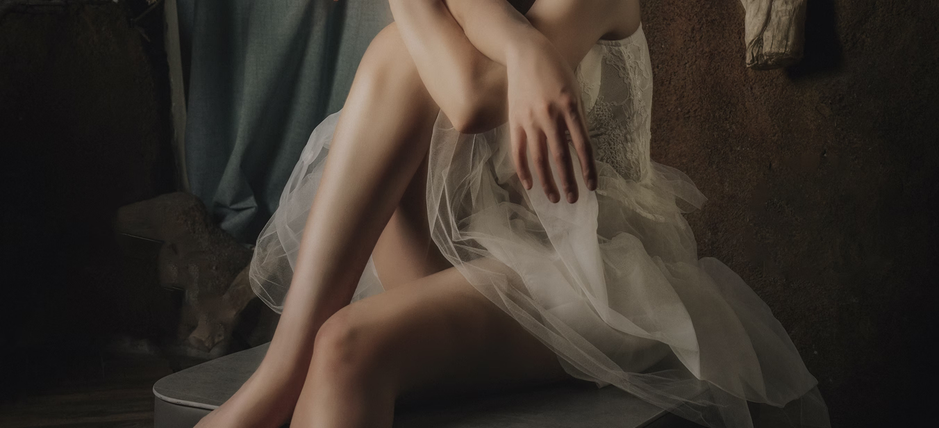 A model sitting with crossed legs, wearing a white tulle dress, with one hand resting on her knee and another on her lap.