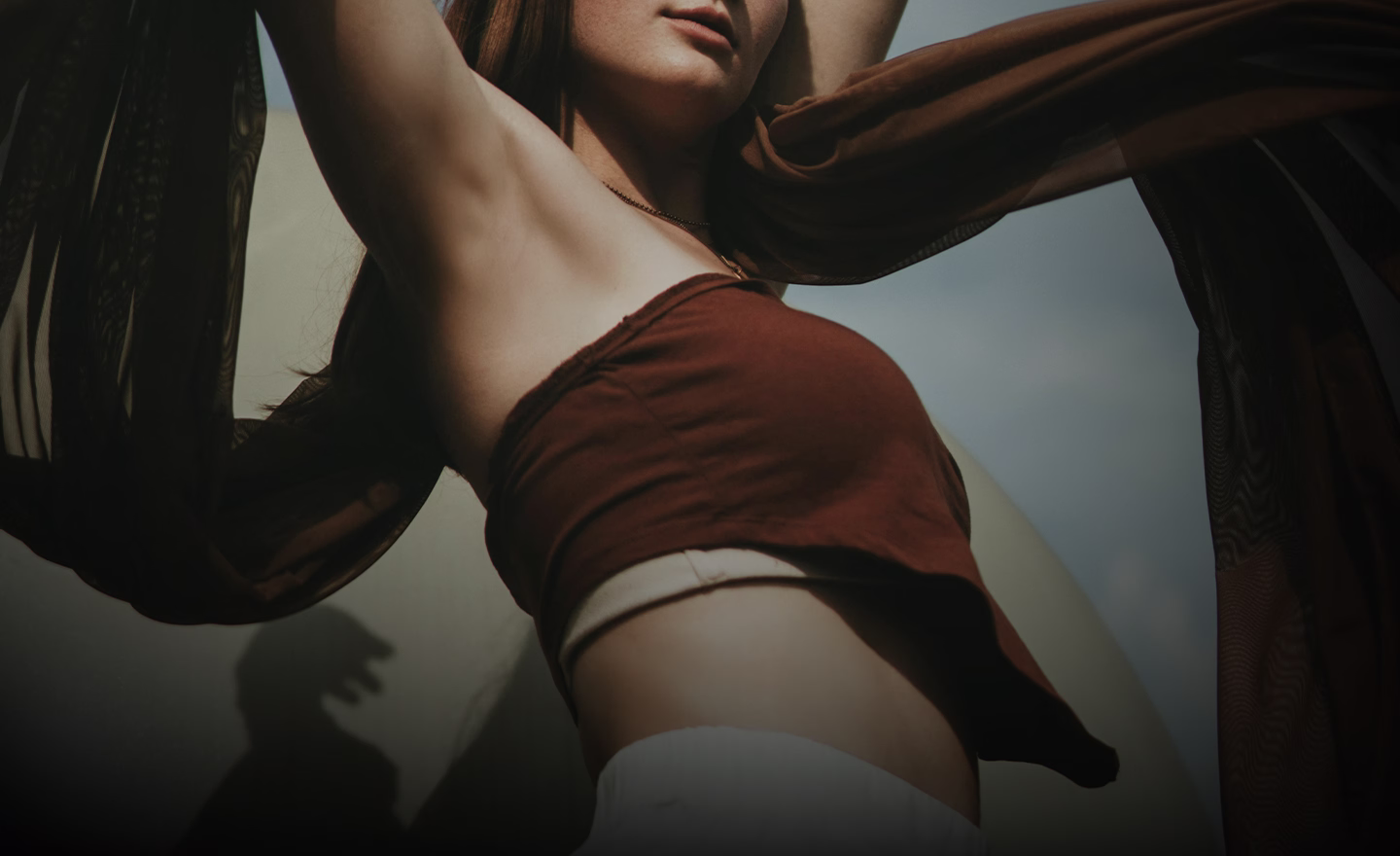 A woman wearing a brown crop top, with her arms raised, holding a flowing scarf.