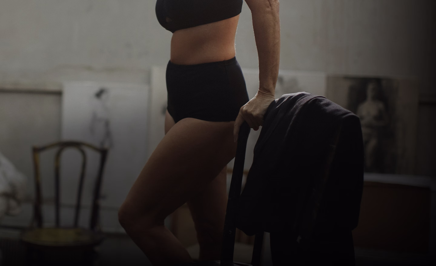 A woman in black high-waisted shorts and a matching cropped top, standing with one hand on a chair and the other holding a black jacket.