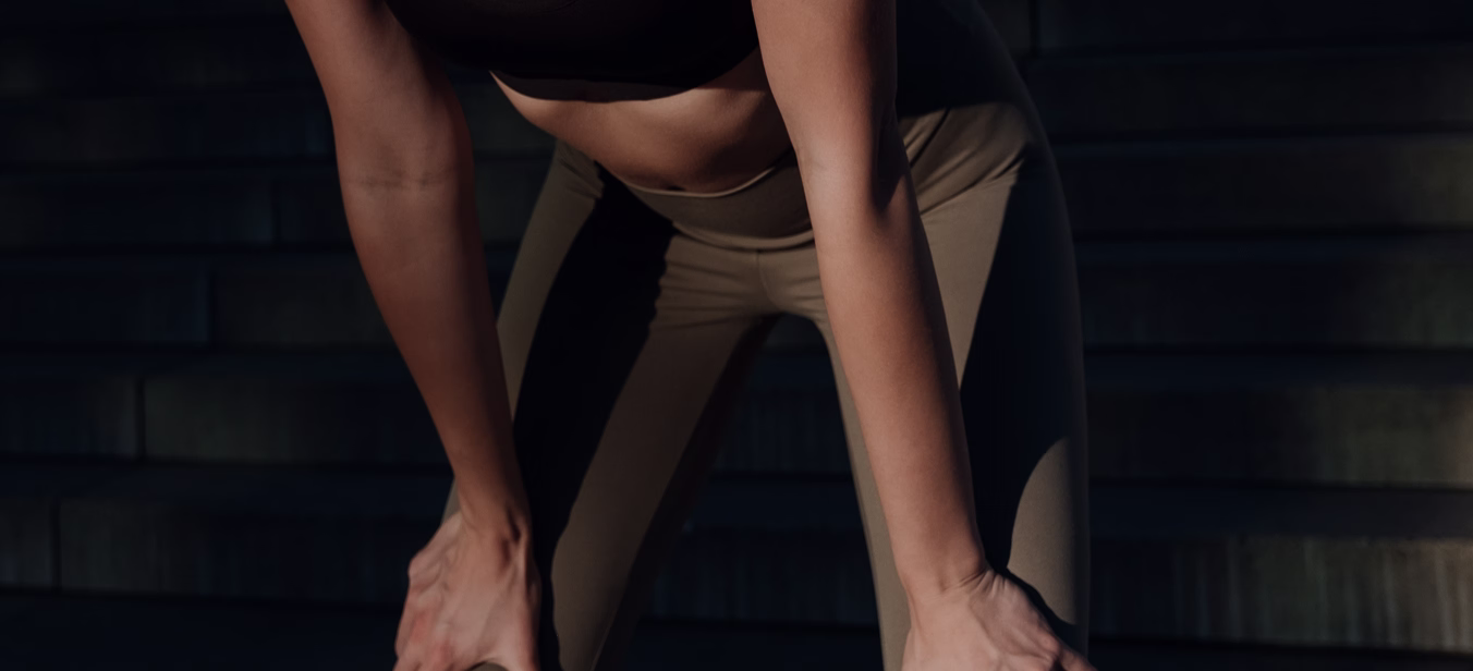 A fit woman wearing a black crop top and beige leggings is crouched down, resting her hands on her knees, with a focused expression.