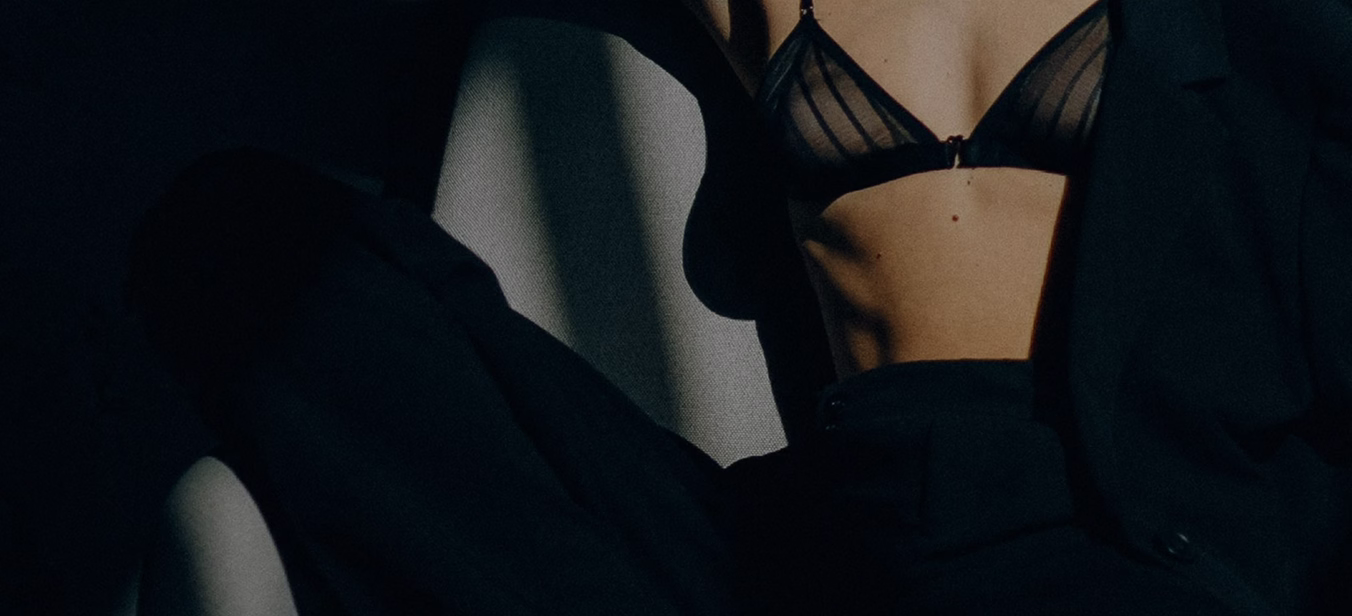A close-up of a person wearing a black sheer bra and black clothing, seated in a relaxed position. The focus is on the torso, highlighting the bra's design and the person's silhouette.