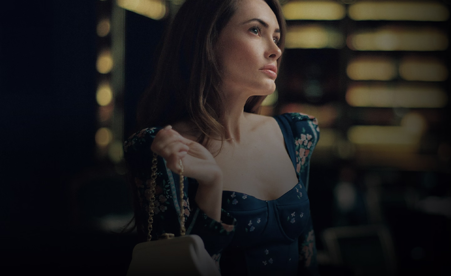 A woman with long brown hair wearing a blue floral dress, holding a white handbag, gazing thoughtfully to her right.