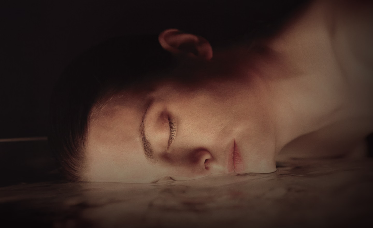A close-up image of a woman lying on a smooth surface with her face turned to the side, eyes closed and expression serene.
