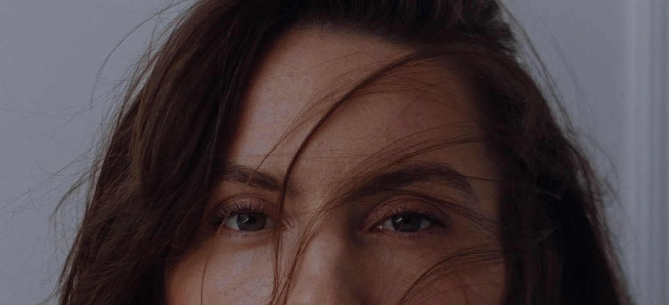 A close-up image of a woman's face with brown hair partially covering her forehead and cheeks, revealing her greenish-brown eyes and natural makeup.