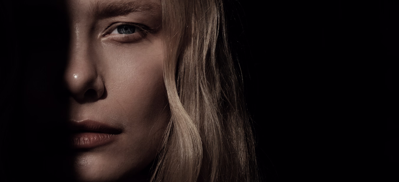 A close-up portrait of a woman with long blonde hair and a serious expression, partially illuminated to highlight her features.