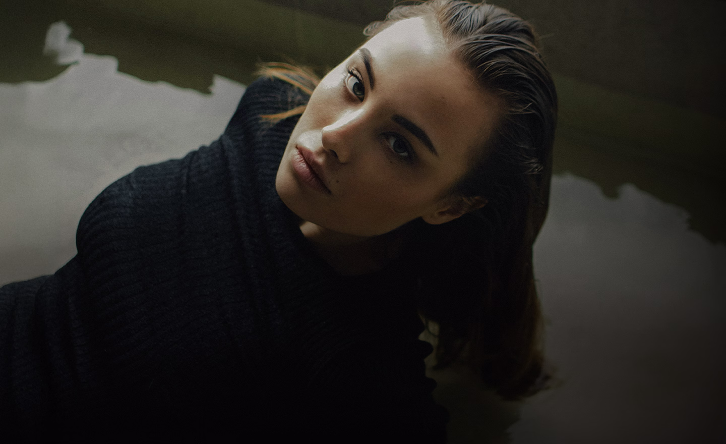 A woman with long brown hair and a serious expression, wearing a black sweater, partially submerged in water.
