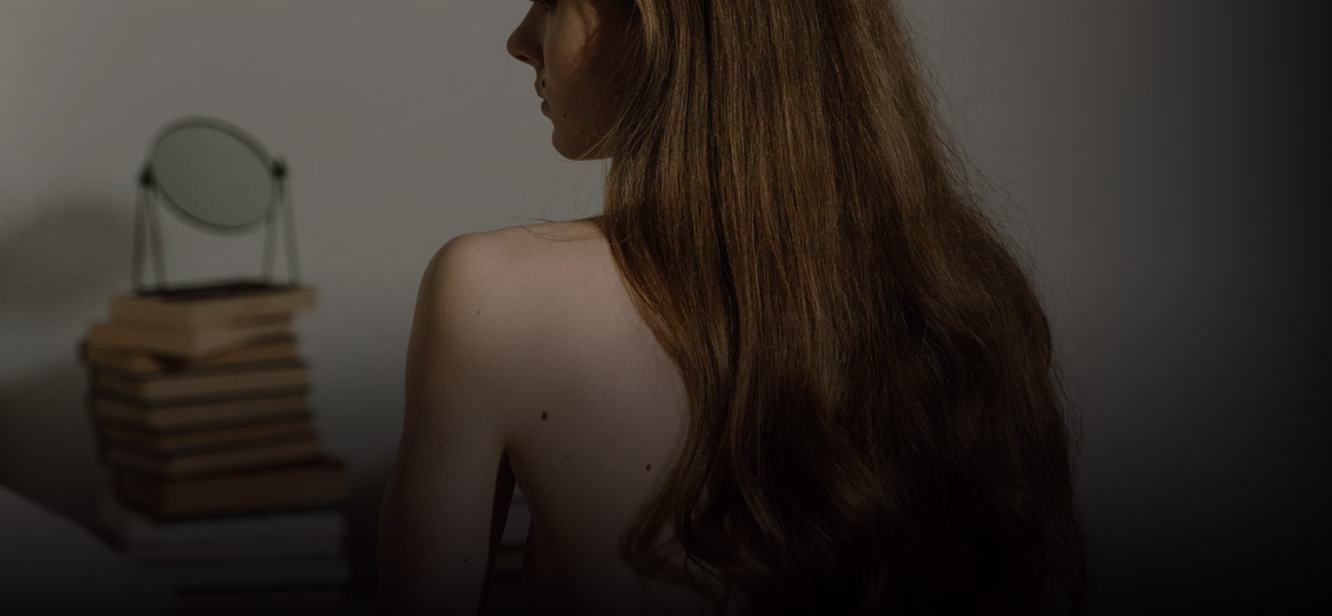 A woman with long brown hair sitting with her back to the viewer, revealing part of her back. A stack of books is visible in the foreground.