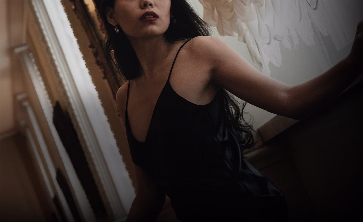 A woman wearing a black sleeveless dress, with long dark hair cascading over her shoulder, posed against an elegant interior setting.