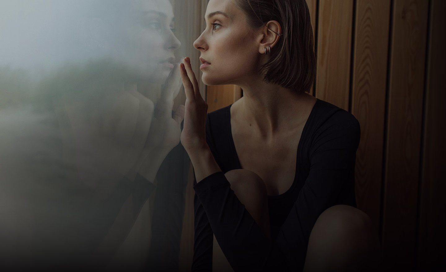 A woman with short brown hair and subtle makeup is contemplatively resting her chin on her hand while gazing out, her profile visible with soft lighting highlighting her facial features.