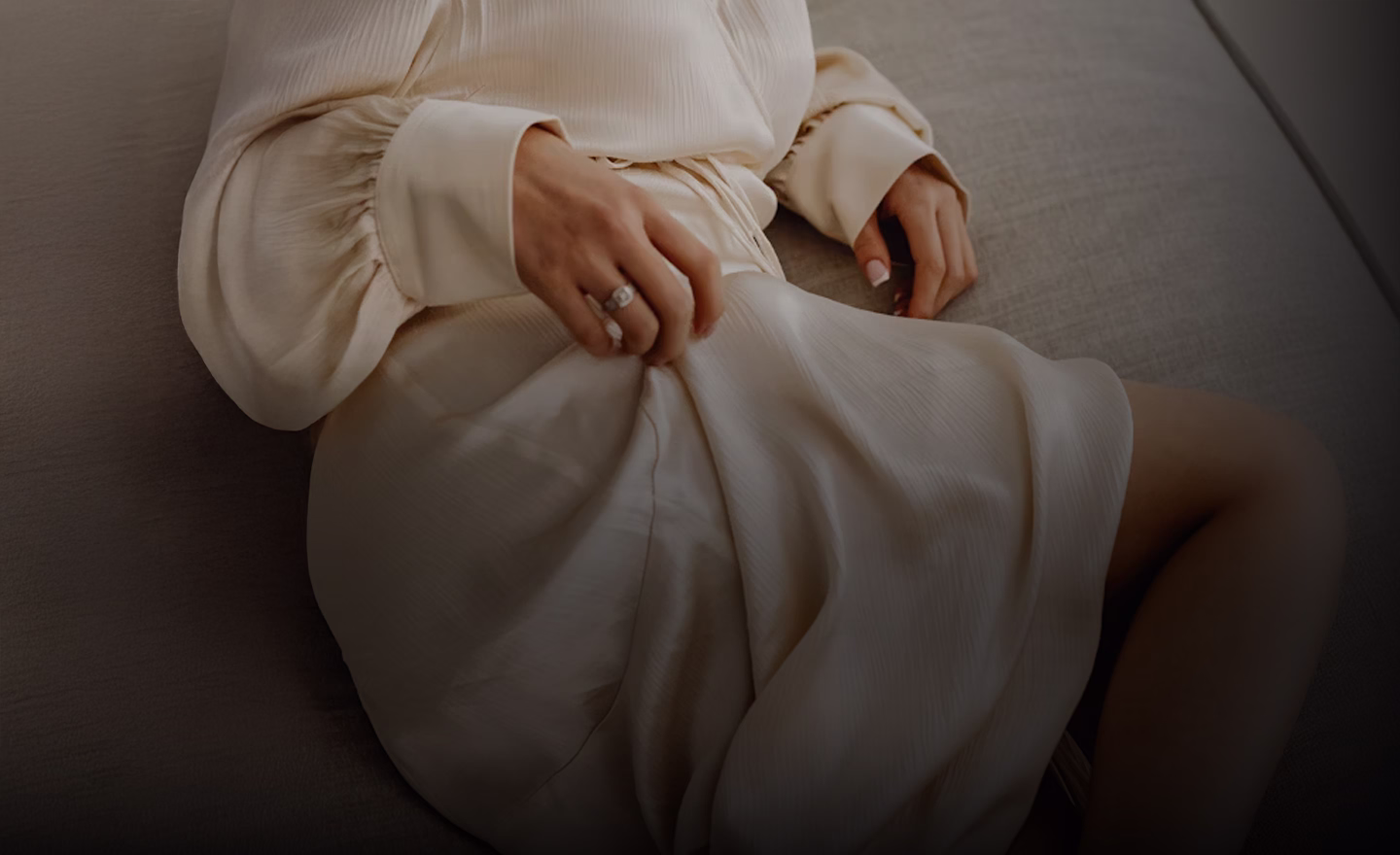 A close-up of a person wearing a light-colored blouse and sitting on a surface, gently adjusting the fabric of a light-colored skirt while wearing a ring on their hand.