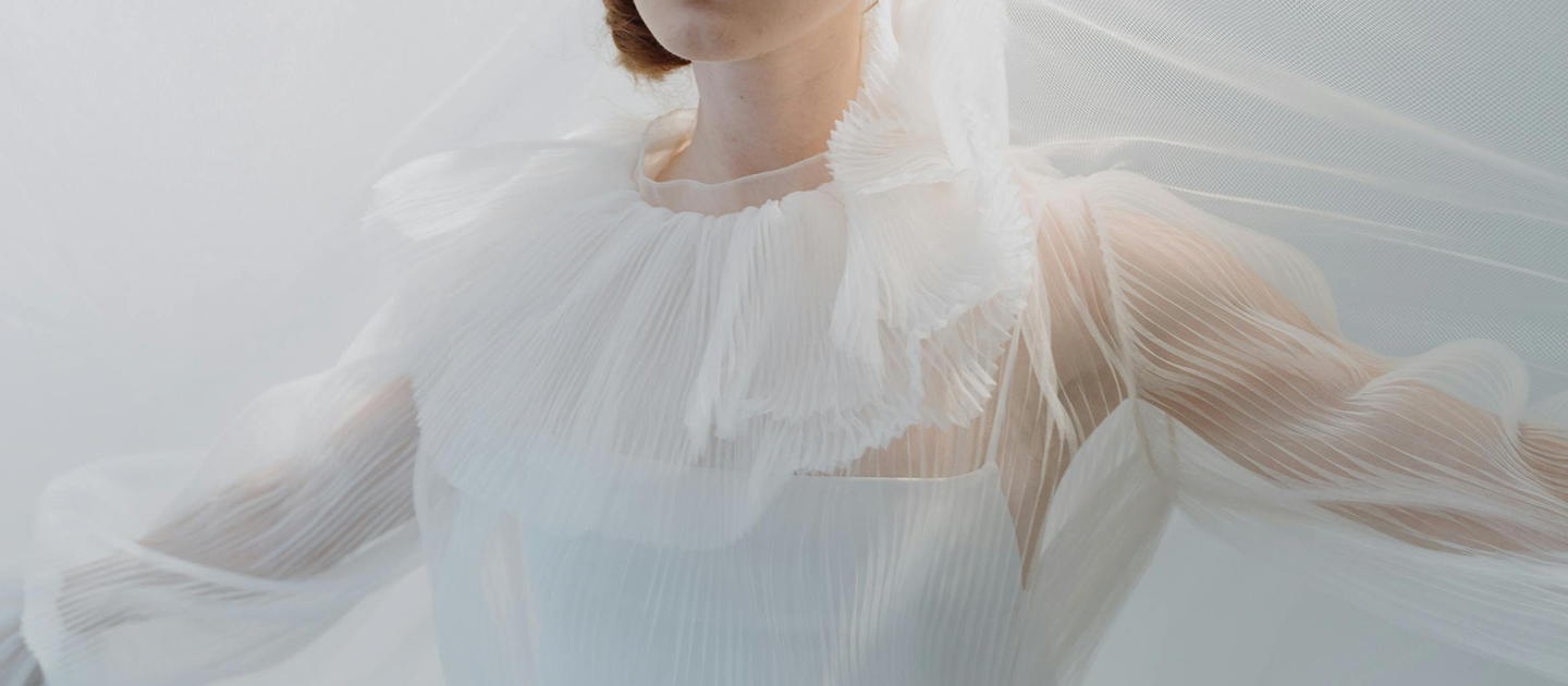 A close-up of a model wearing a sheer, pleated white dress with a ruffled collar. The dress has long, flowing sleeves, and the model's hair is styled in a simple updo.