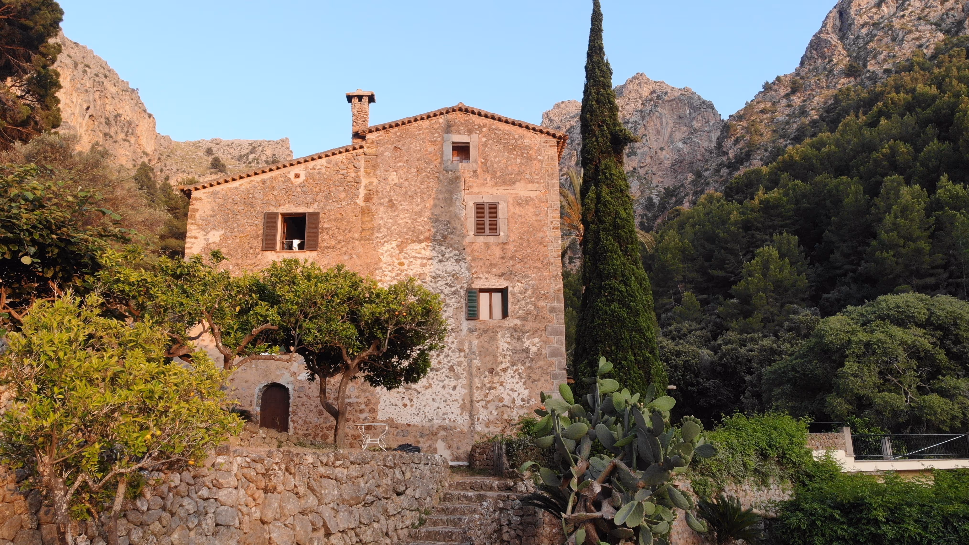 Rustic stone facade of Finca Higo with cypress tree and mountain backdrop, nestled in the Tramuntana mountains.