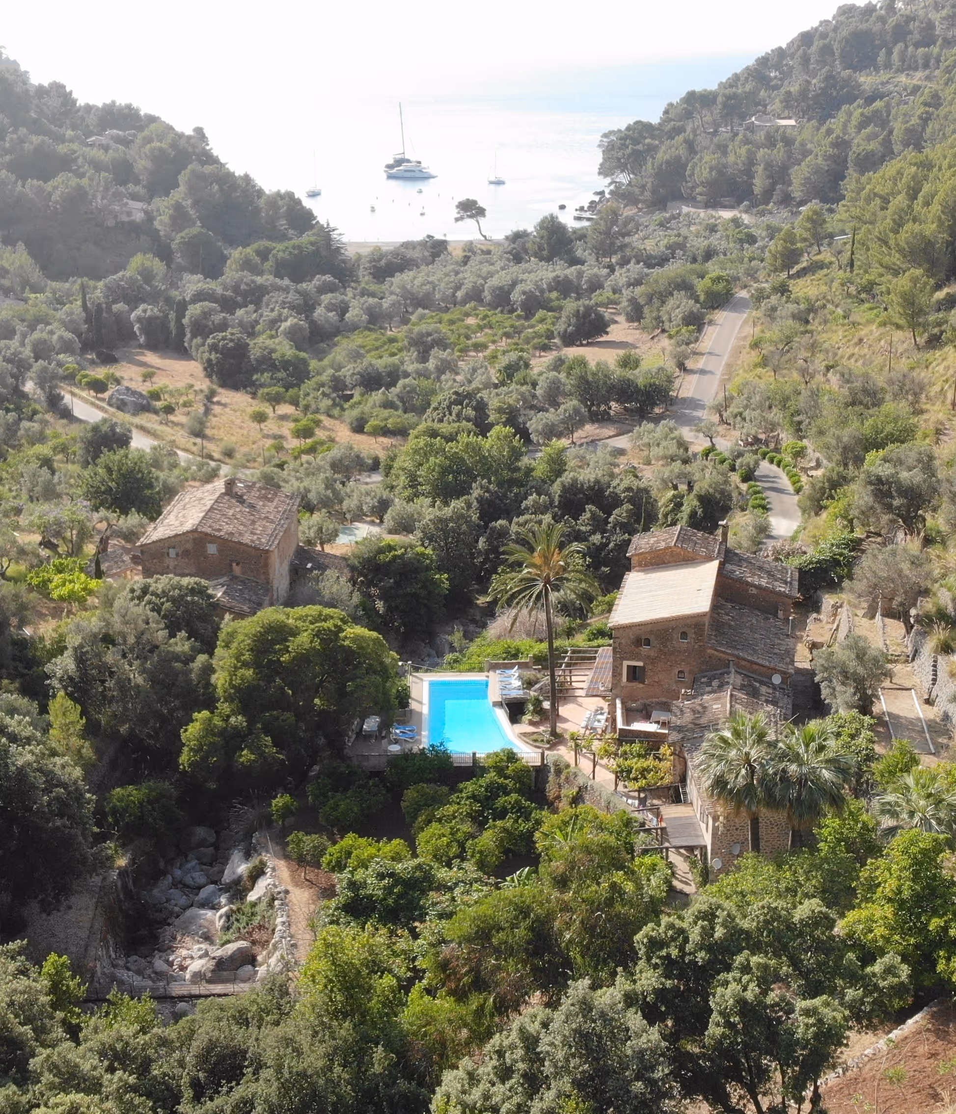 Aerial view of Finca Higo surrounded by lush greenery and mountains, with a pool in the center and a scenic coastal bay with anchored boats in the background.