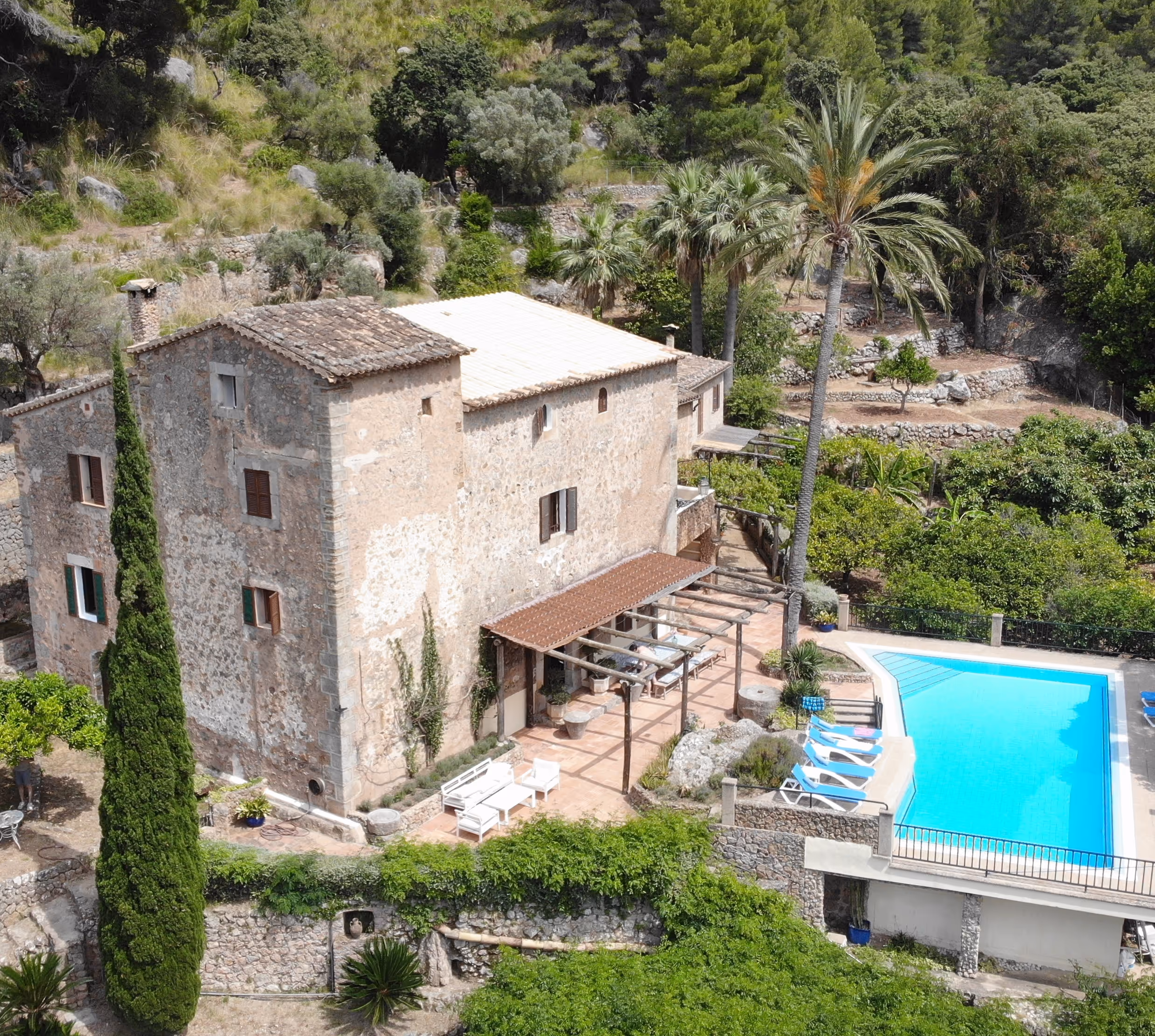 Aerial view of Finca Higo’s traditional stone house with a shaded terrace, sun loungers, and a bright blue pool surrounded by lush greenery and palm trees in the Tramuntana mountains.