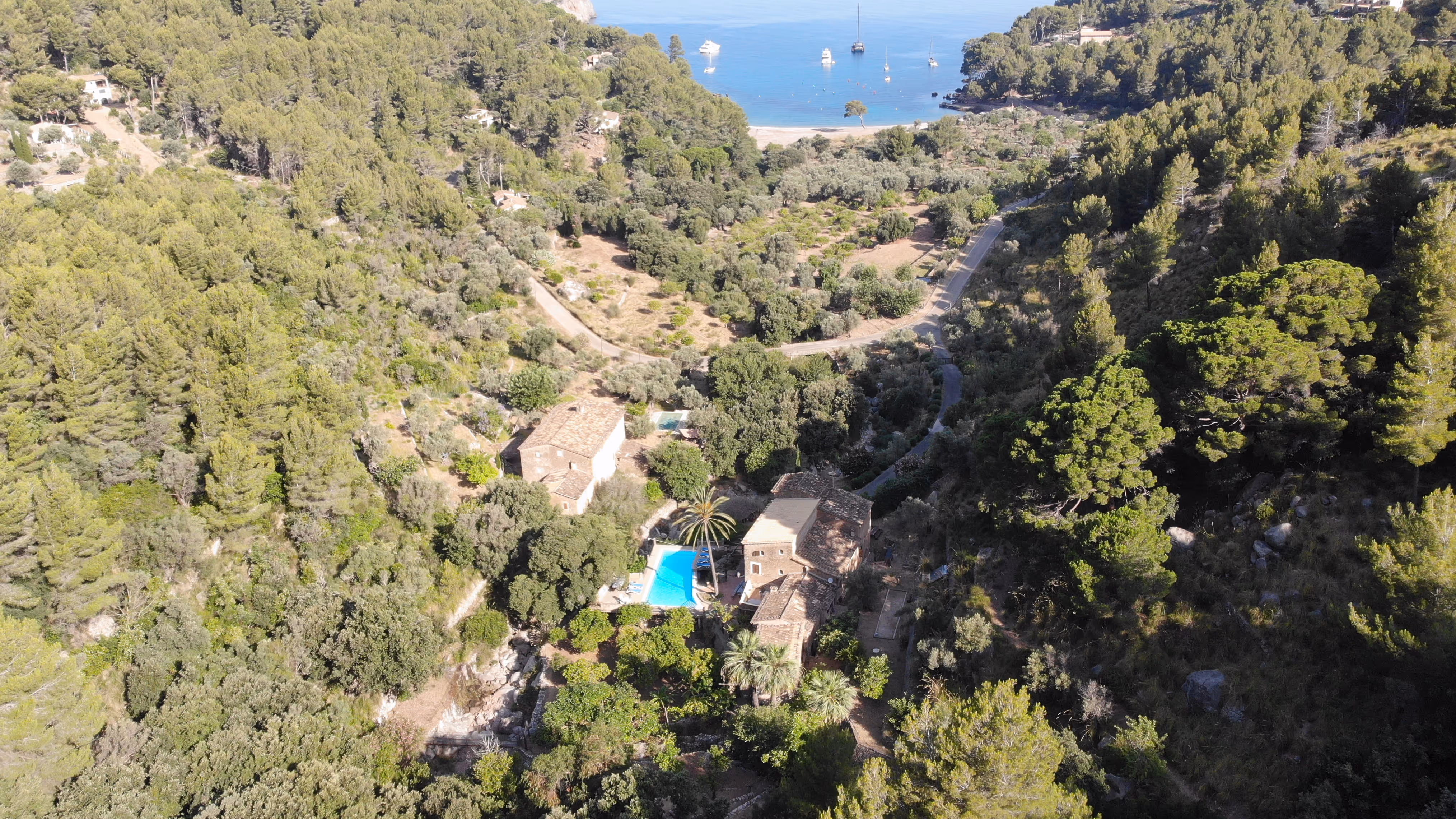 Aerial view of Finca Higo retreat nestled in a lush green valley near a tranquil beach with anchored sailboats, surrounded by pine forests in Mallorca