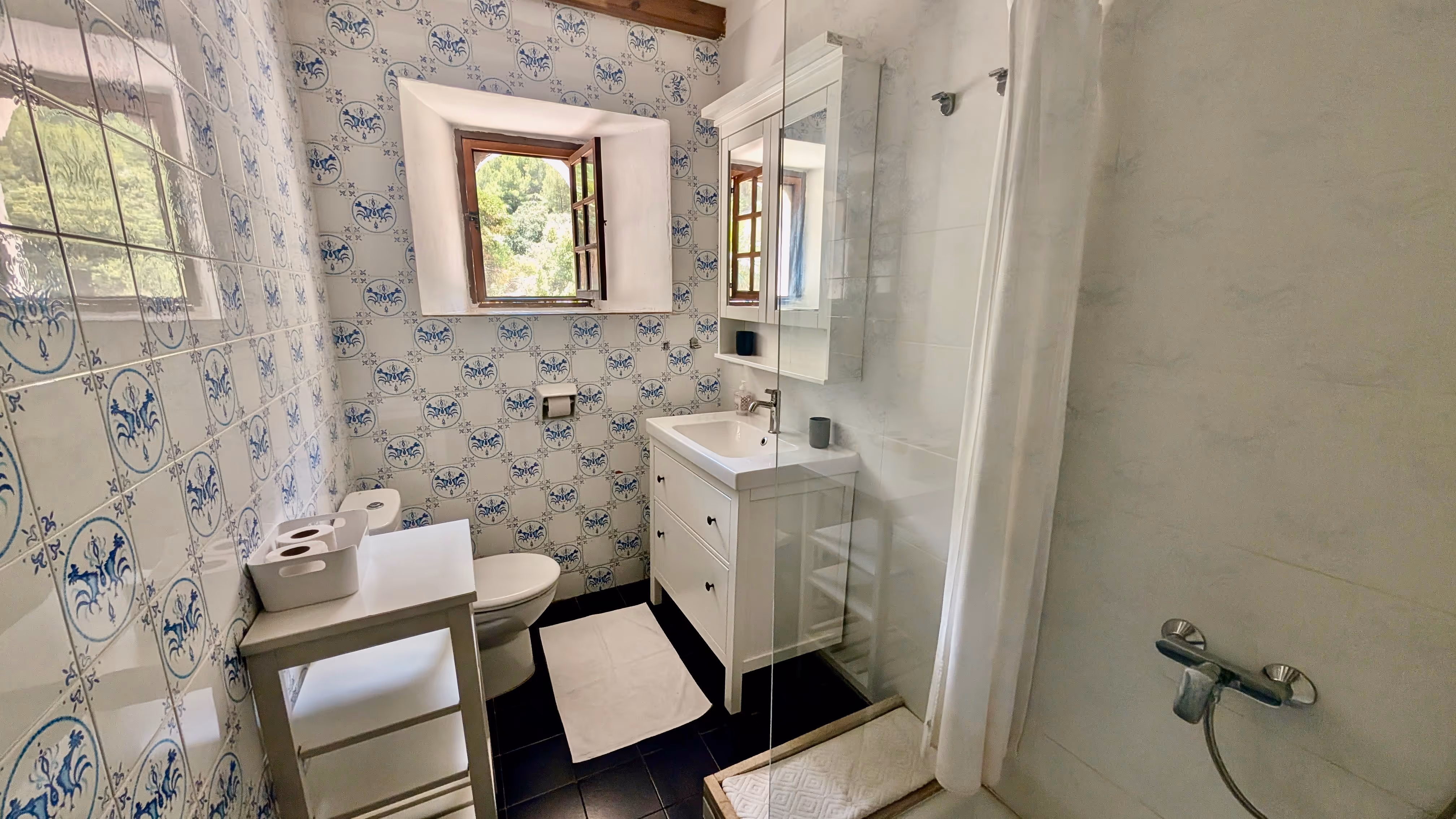 Bright bathroom with patterned tiles, glass shower, and a window looking out to nature.