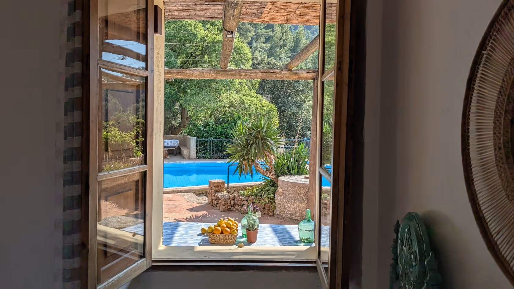 View through an open window at Finca Higo looking out onto a sunlit terrace with a pool, palm trees, and a table set with lemons and water.