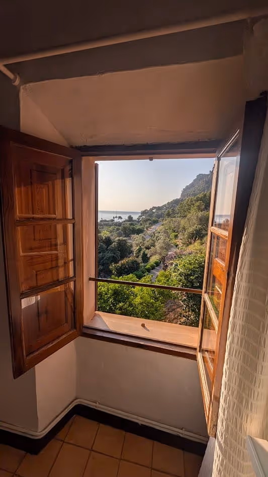 Open wooden window with a scenic view of forested hills leading to the coastline and anchored boats at Cala Tuent.