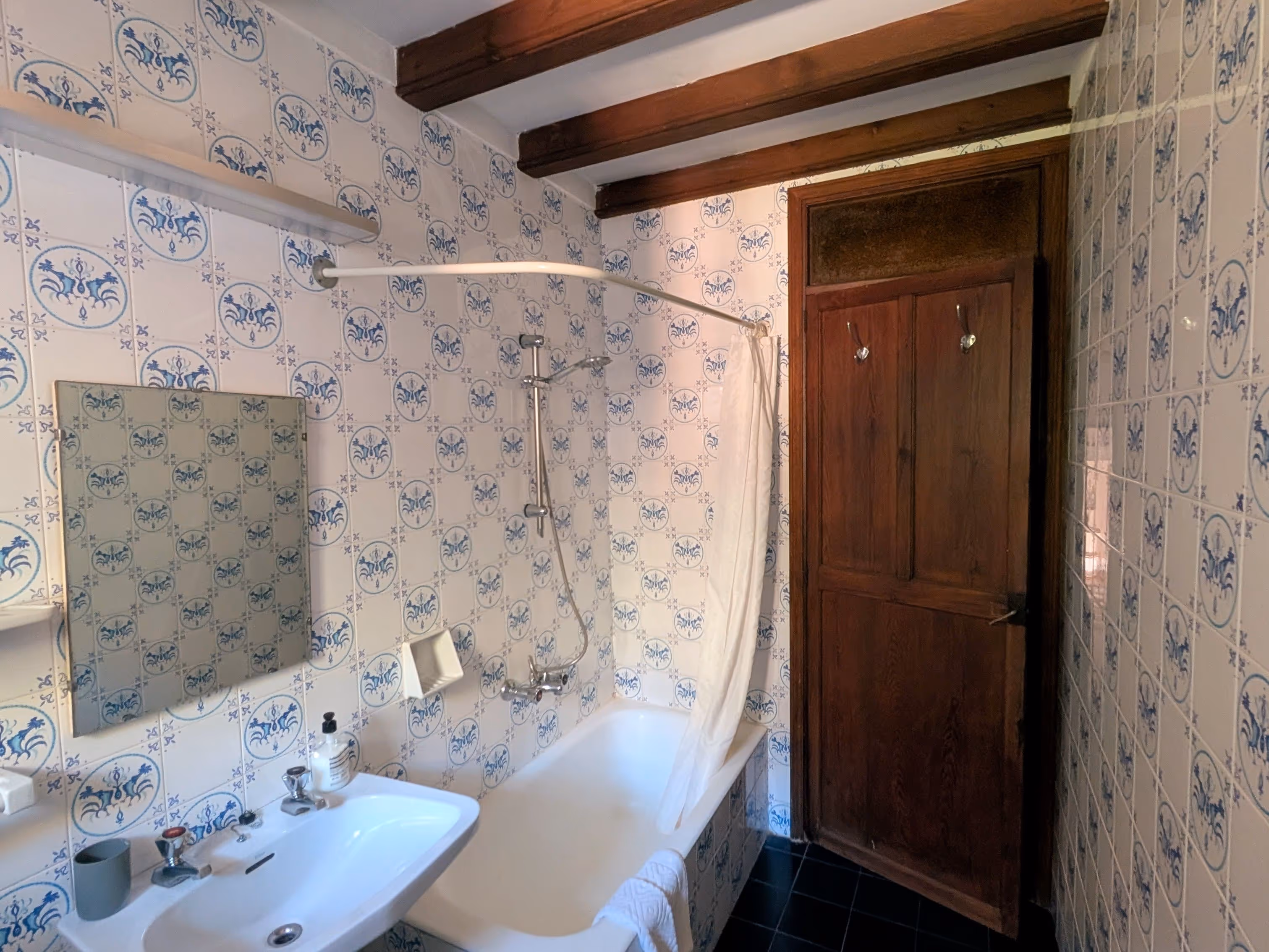 Traditional bathroom with blue-and-white tiled walls, a bathtub with shower curtain, wooden ceiling beams, and a wooden door.