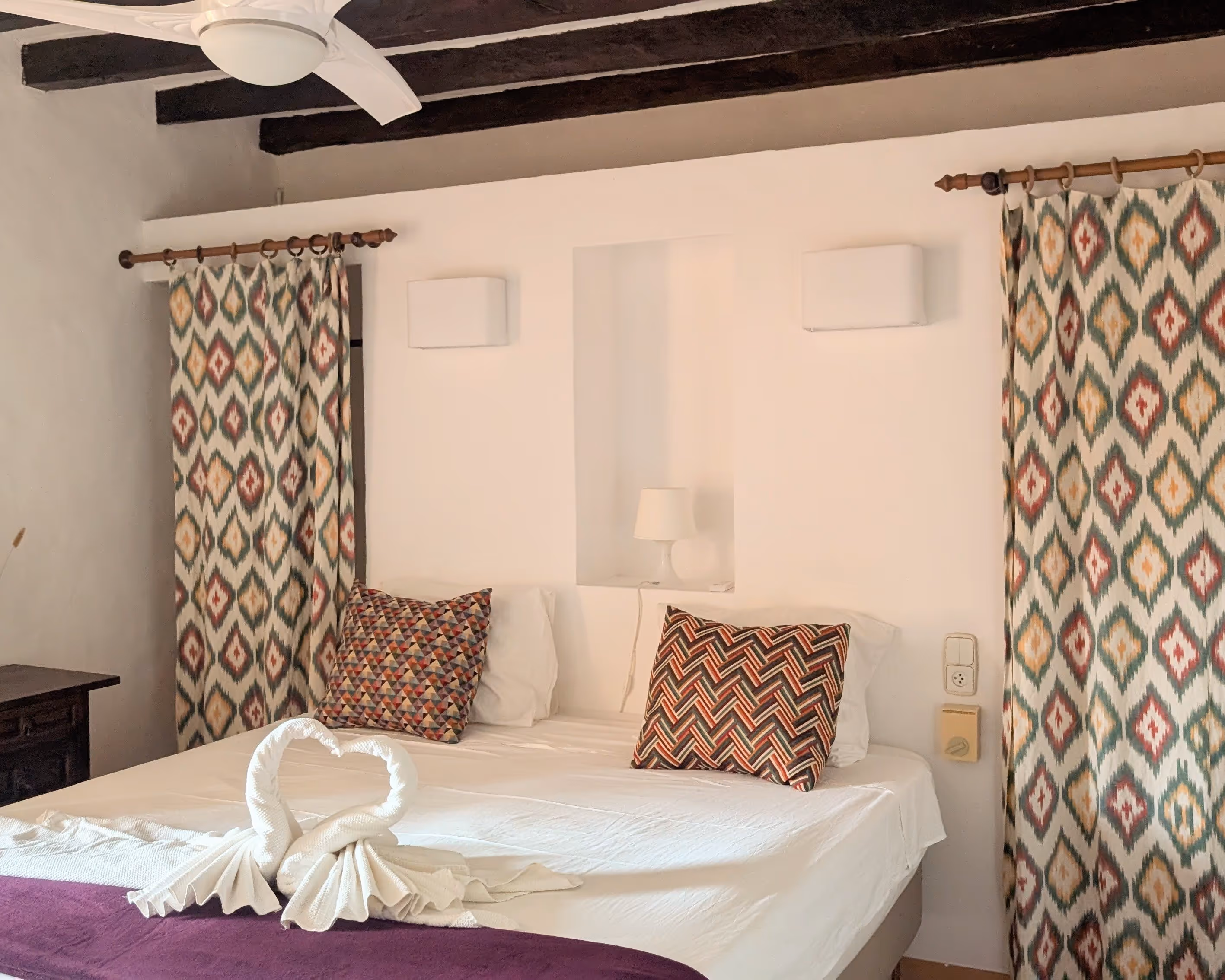 Bright double room with traditional patterned curtains, rustic wooden beams, and a neatly made bed.