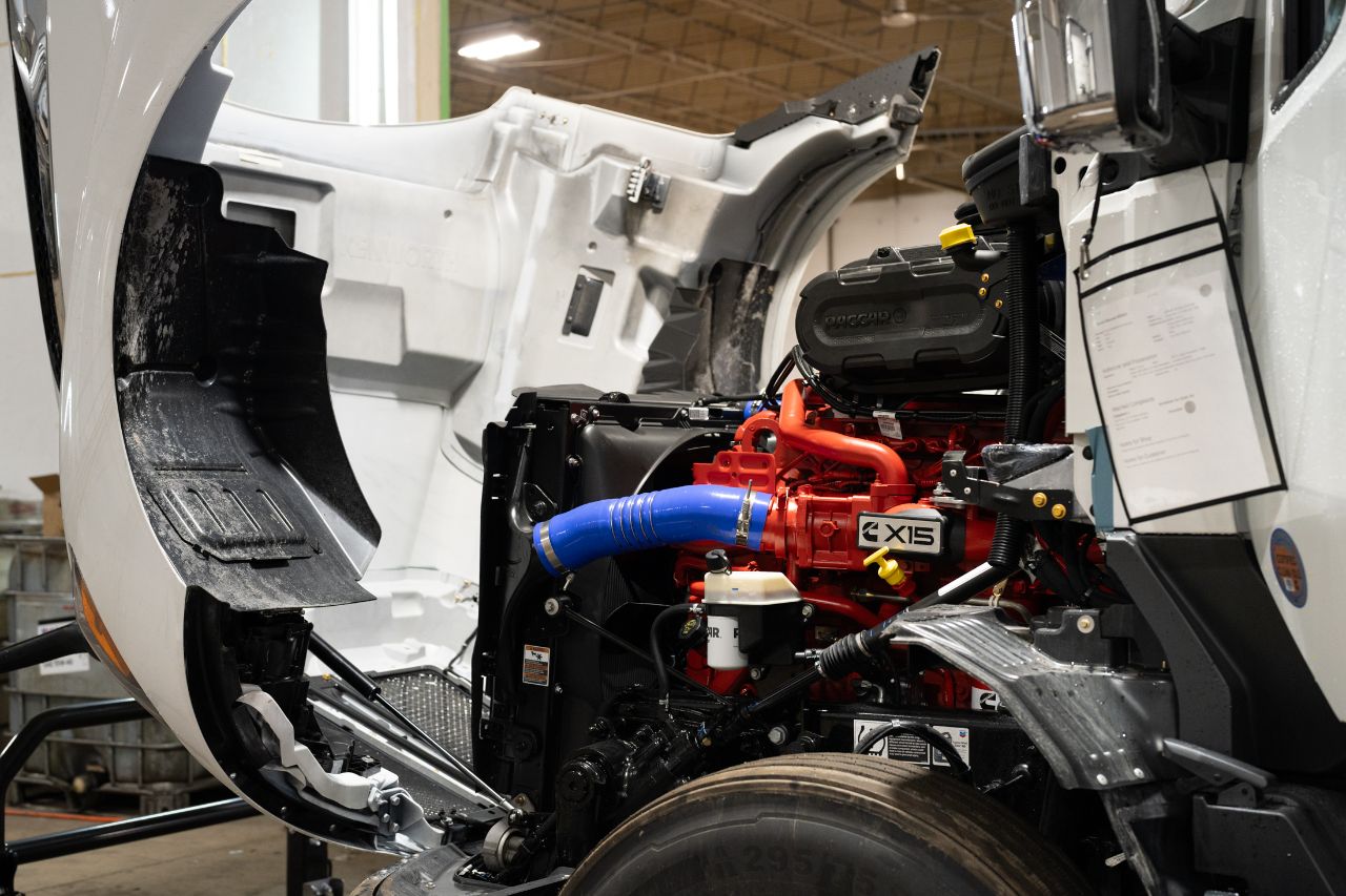 Open hood of a white semi truck revealing a clean Cummins X15 diesel engine with red block and blue intake hose.