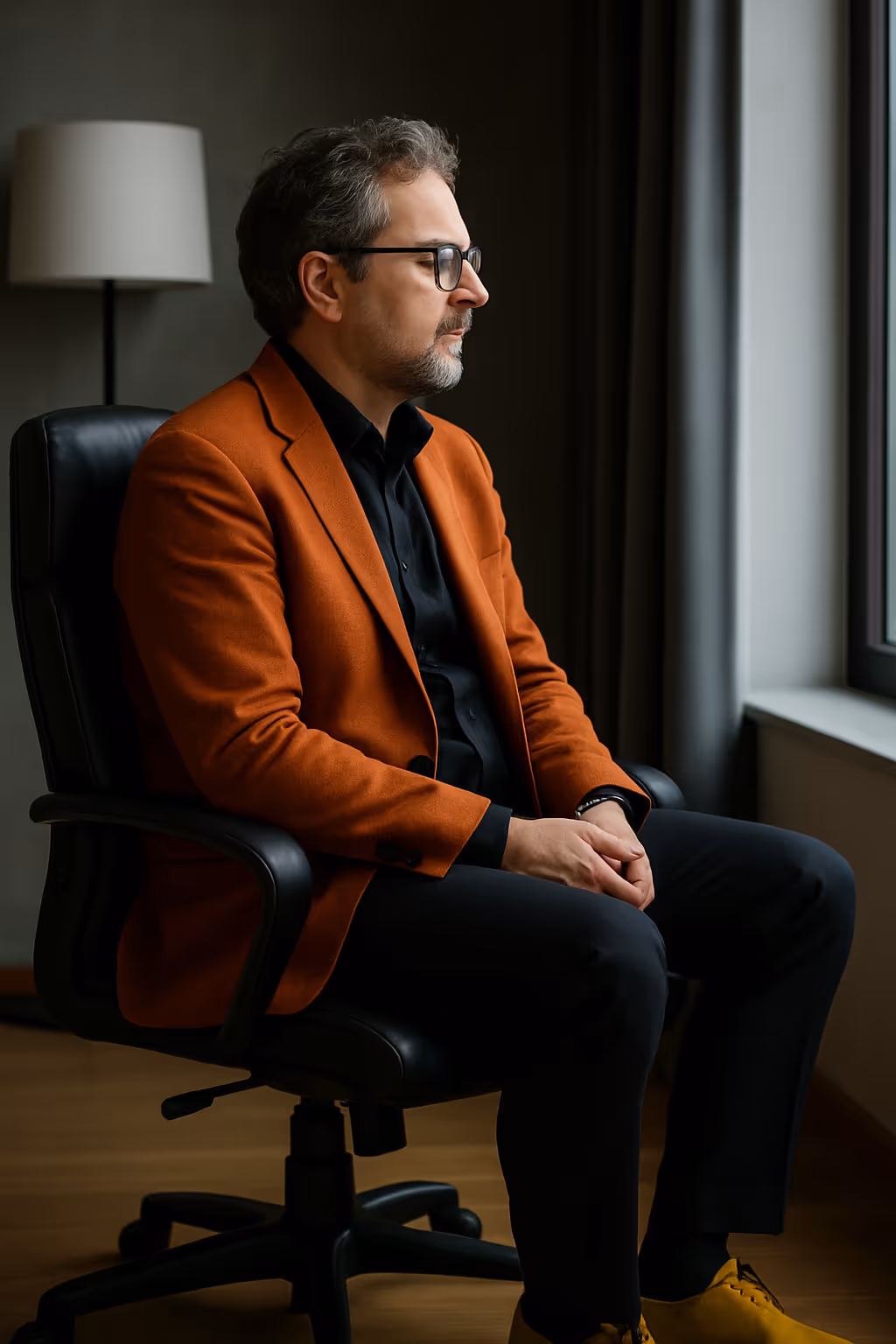 Person sitzt seitlich am Fenster, trägt orangen Blazer und schwarze Kleidung, wirkt nachdenklich.

