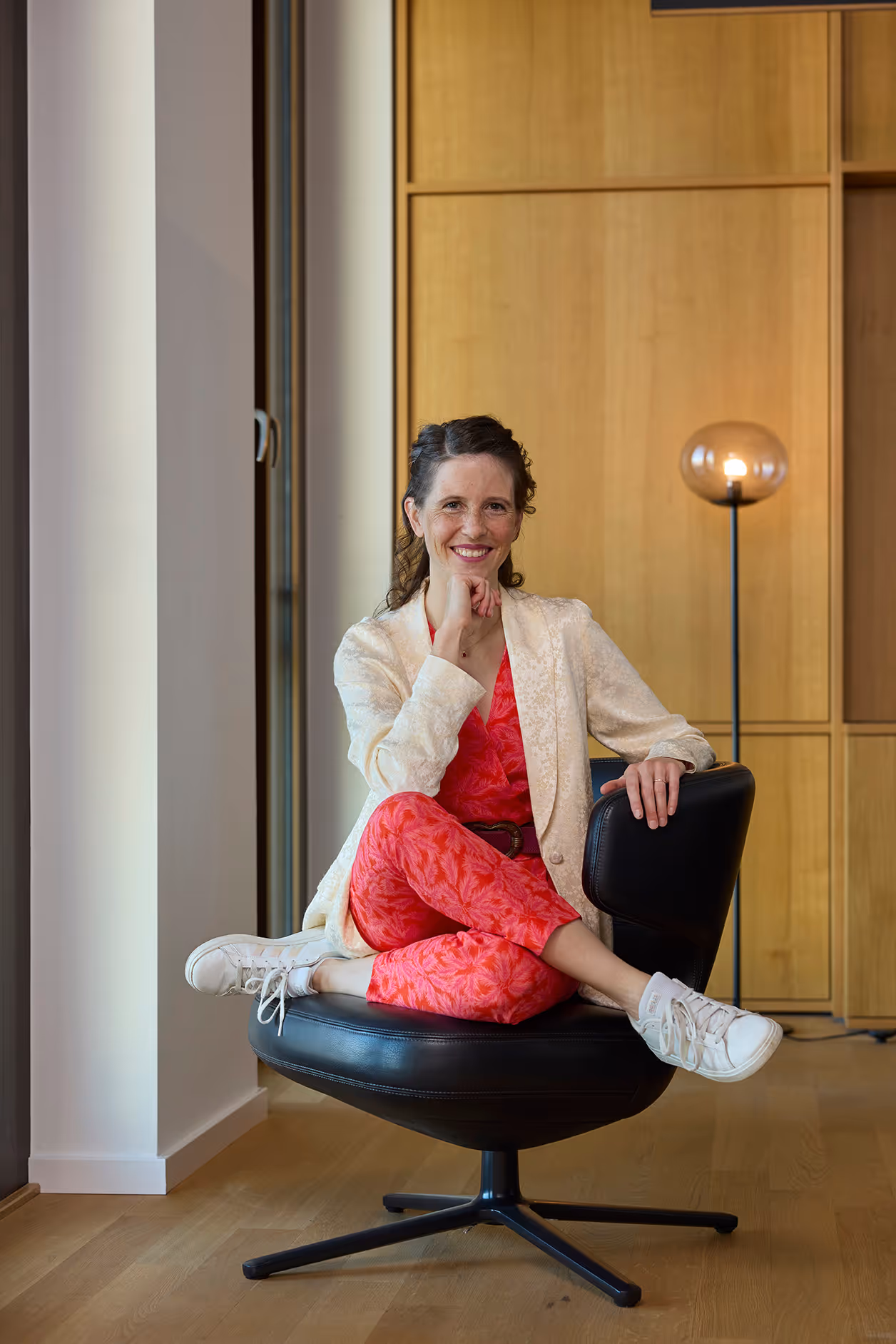 Frau sitzt lächelnd im Sessel, trägt roten Anzug und cremefarbenes Jackett.
