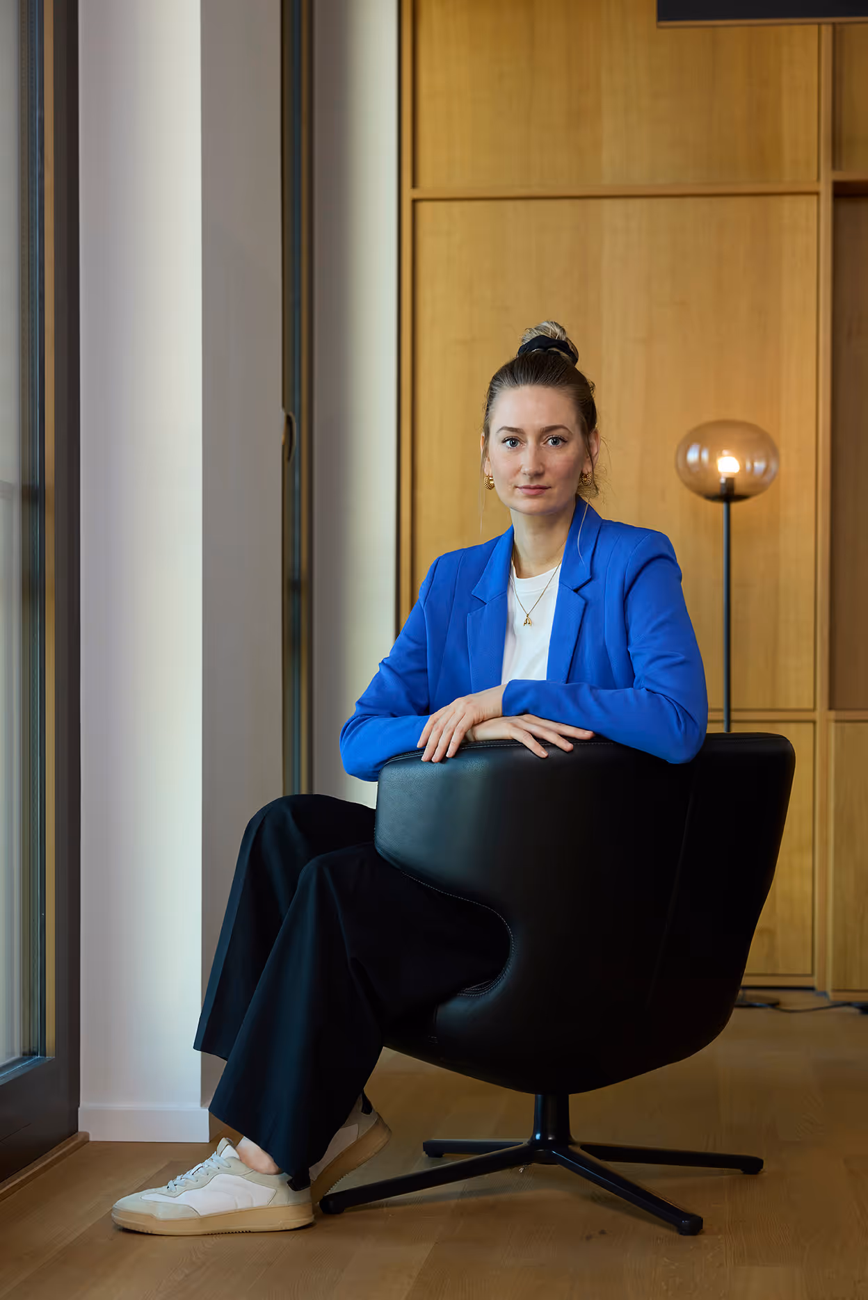 Frau sitzt im schwarzen Sessel, trägt blauen Blazer und schaut direkt in die Kamera.
