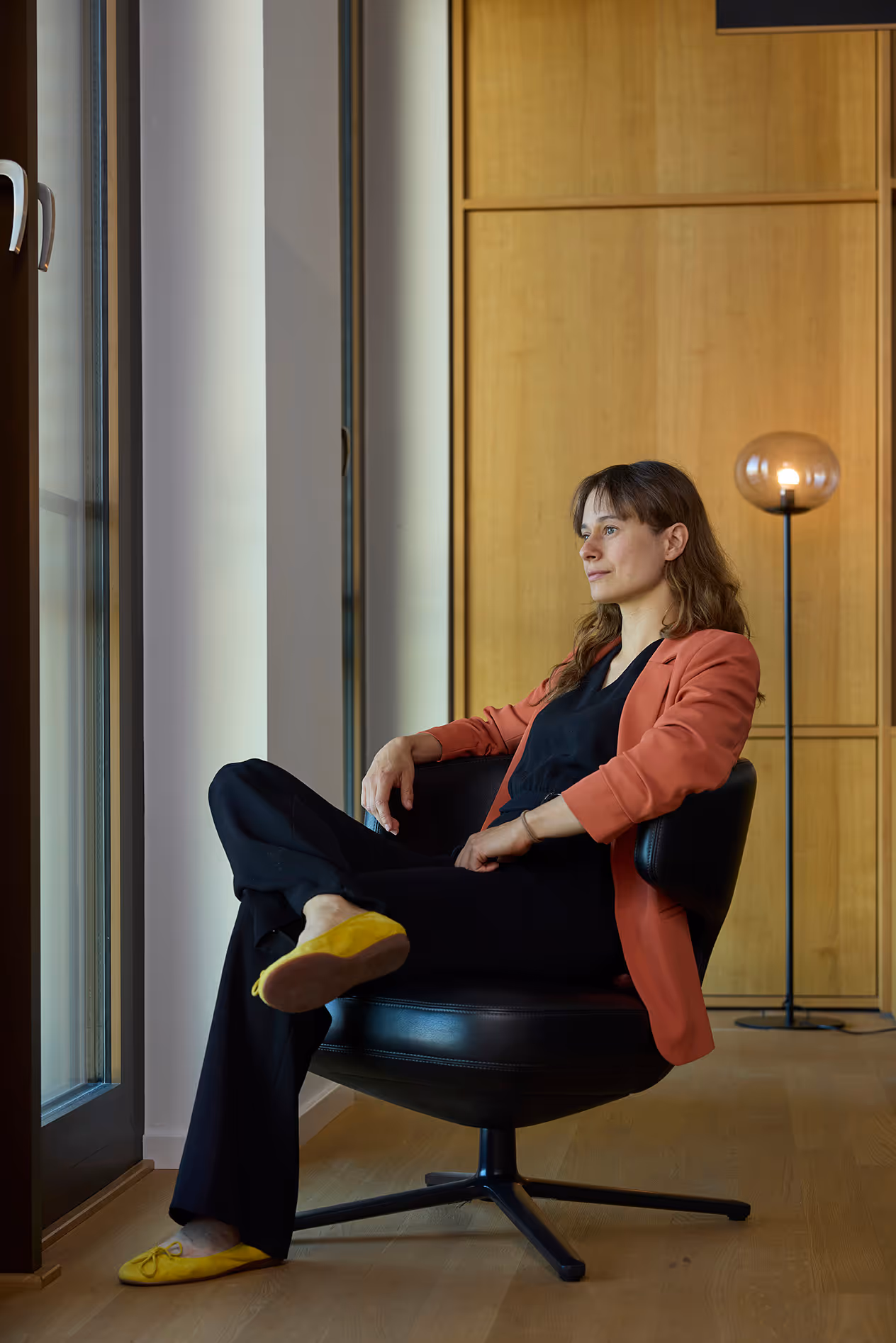 Frau lehnt entspannt im Sessel, trägt rostfarbenes Jackett und gelbe Schuhe.
