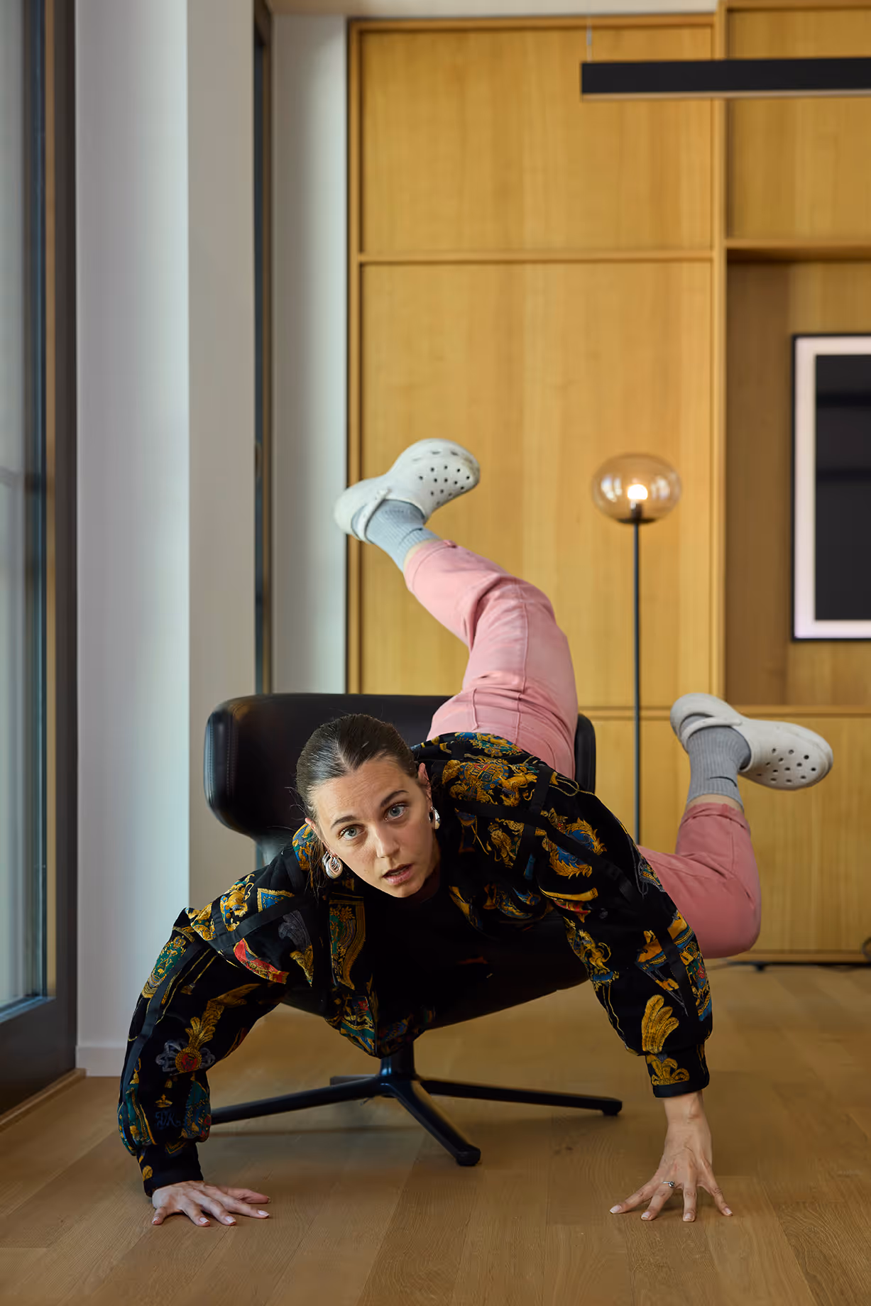 Frau posiert akrobatisch neben Sessel, trägt bunte Jacke und rosa Hose.
