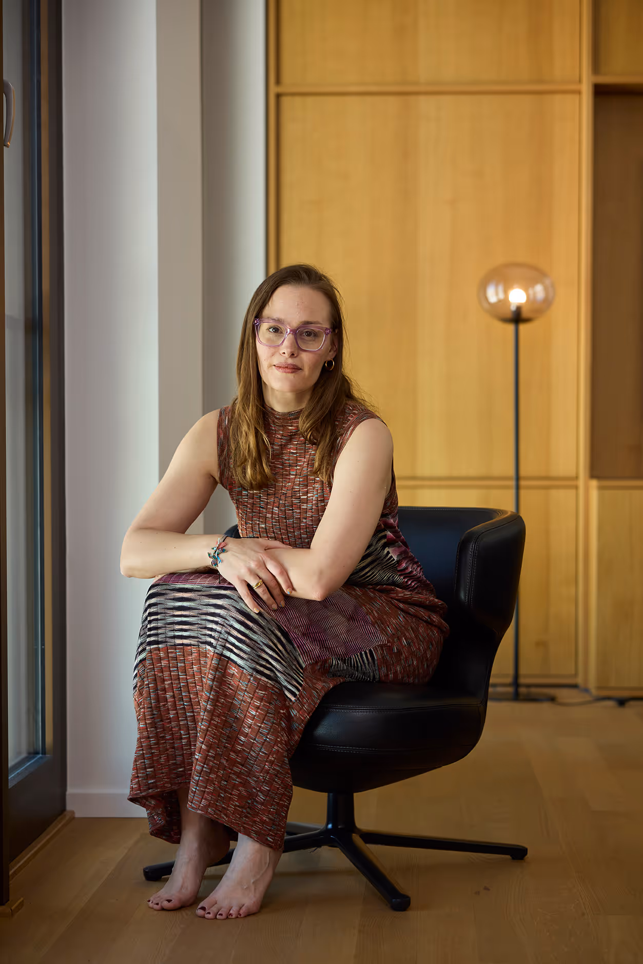 Frau sitzt barfuß im Sessel, trägt ein bunt gemustertes Kleid und Brille.
