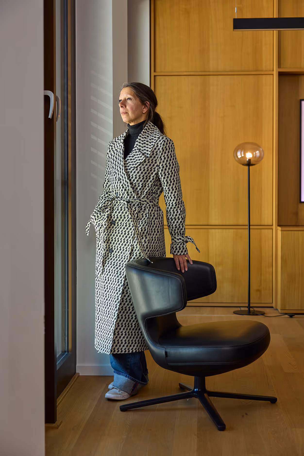 Frau steht neben Sessel, blickt nach draußen und trägt gemusterten Mantel.
