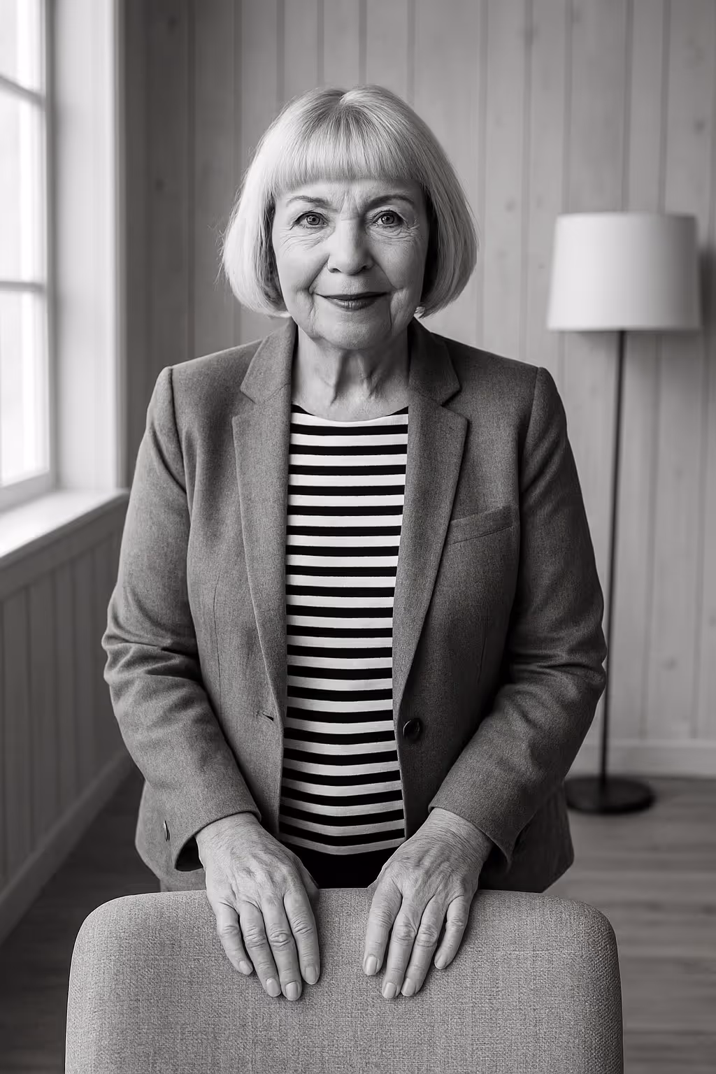 Person steht hinter Stuhl, trägt gestreiftes Shirt und Blazer, Porträt in Schwarz-Weiß.
