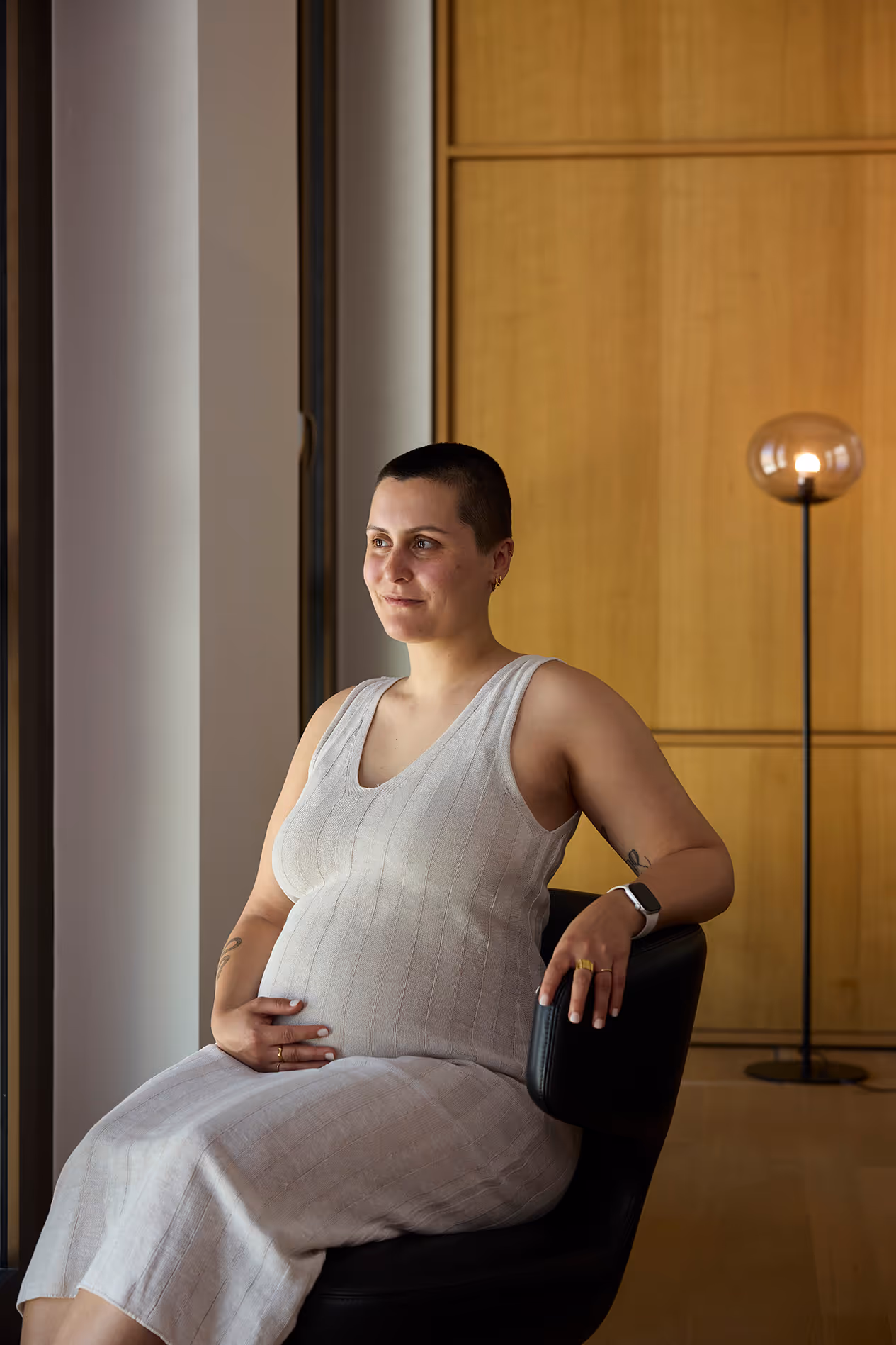 Schwangere Person sitzt im Sessel, trägt helles Kleid und blickt ruhig aus dem Fenster.
